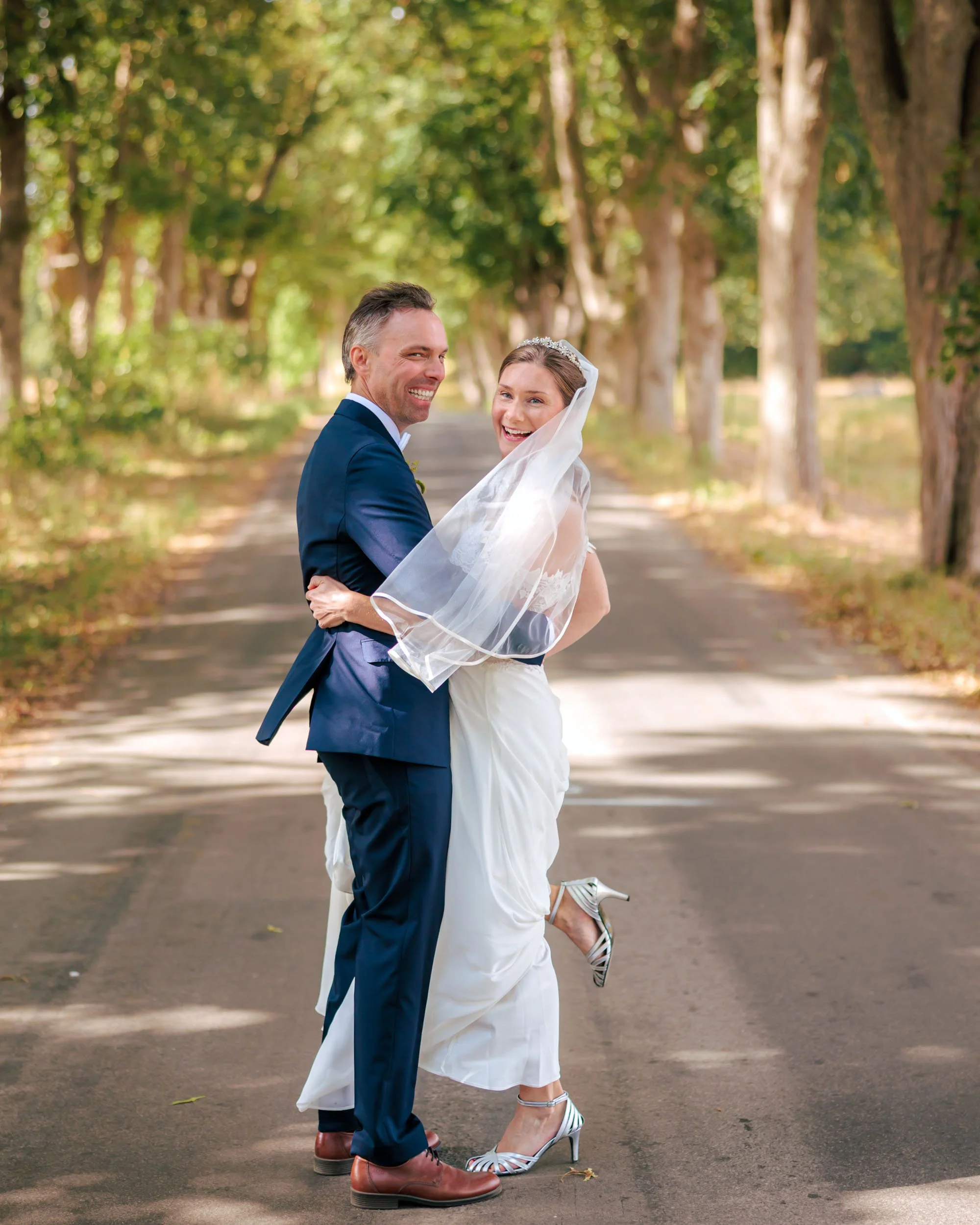 Nytt gift par på en trädgårdsväg, han i mörk kostym och hon i brudklänning och slöja ler mot kameran.
