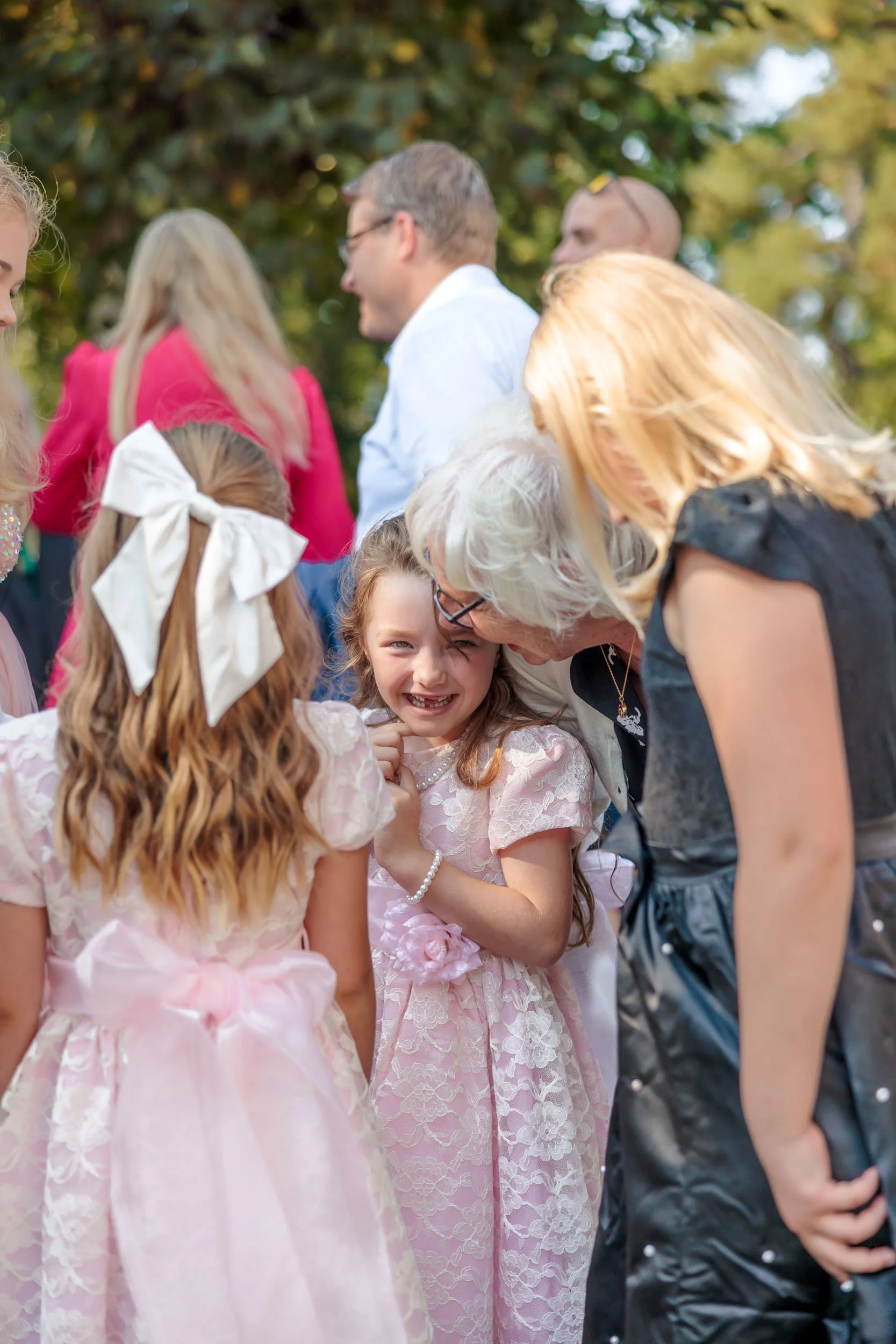 En grupp människor, inklusive en ung flicka i rosa festklänning som ler och pratar med en äldre kvinna, utomhus på en höstdag.
