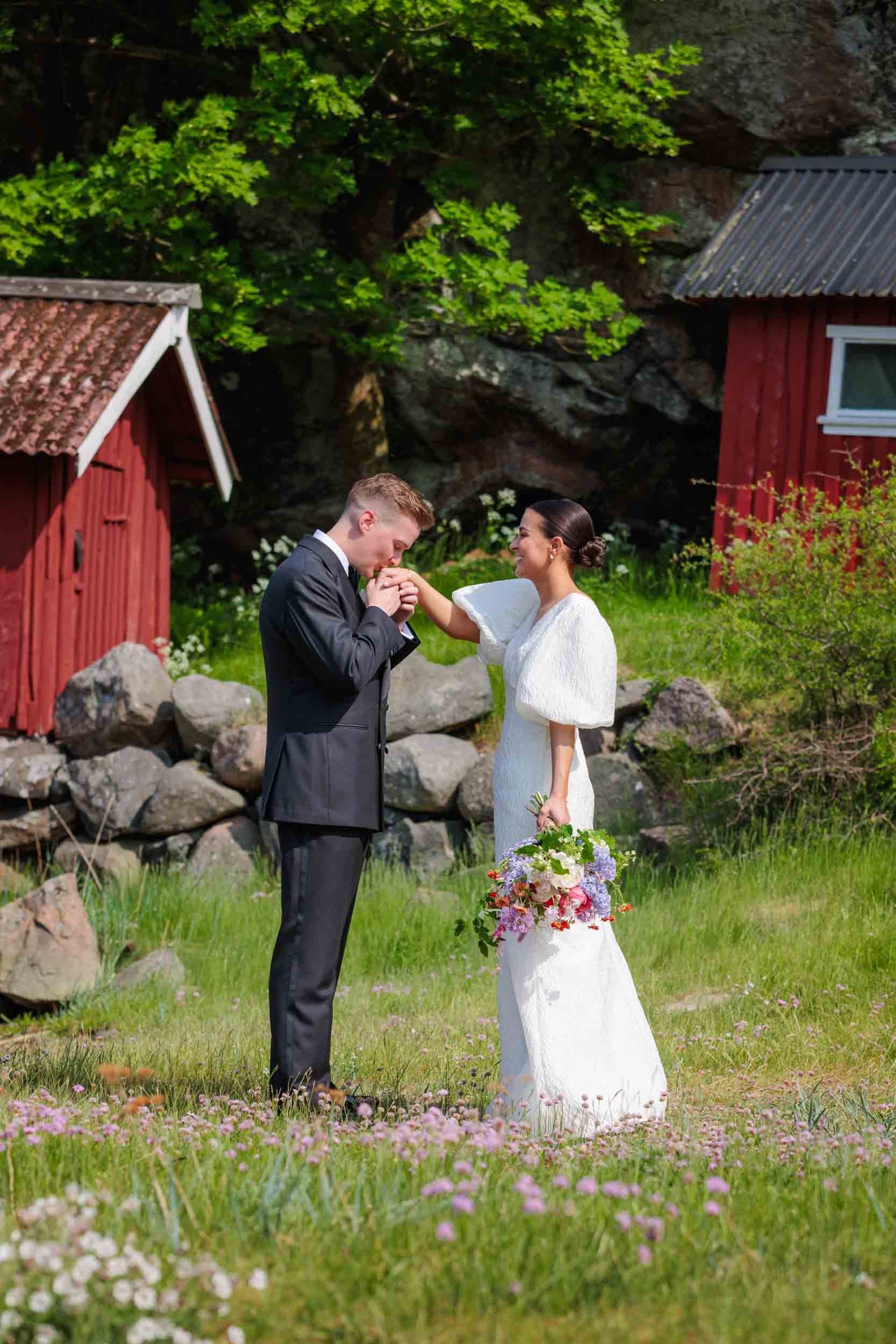 Brudgum kysser brudens hand i naturlig stund framför stenmur och röd lada i sommargrönt