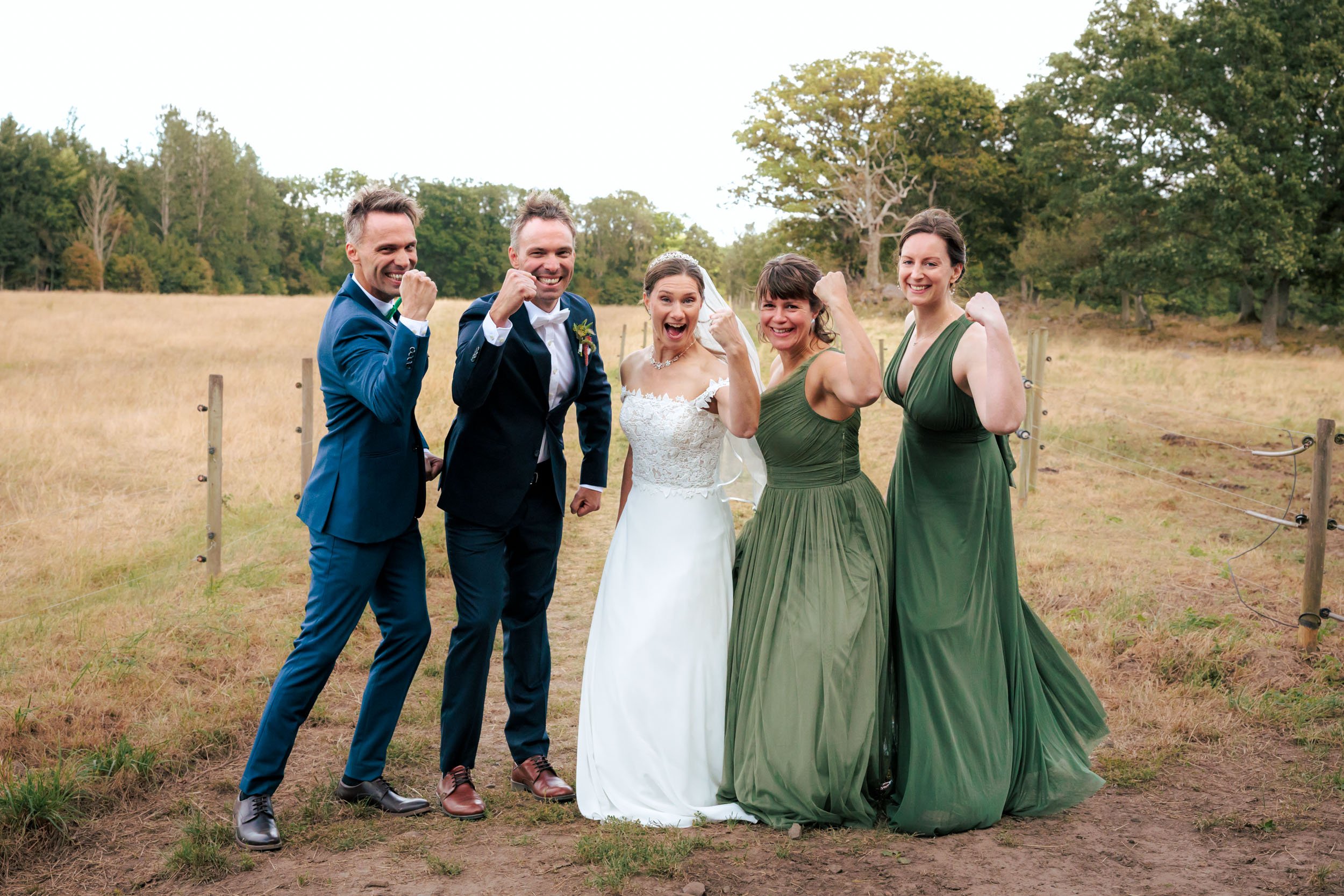 Fem personer, tre män och två kvinnor, poserar utomhus under en bröllopsfest, alla med armarna i flexposition