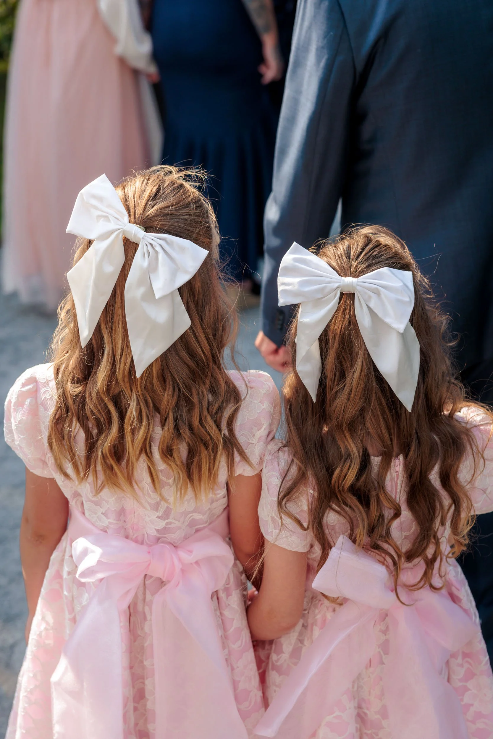 Två flickor i ljusrosa blommande klänningar med stora vita rosetter i håret, sedd bakifrån.
