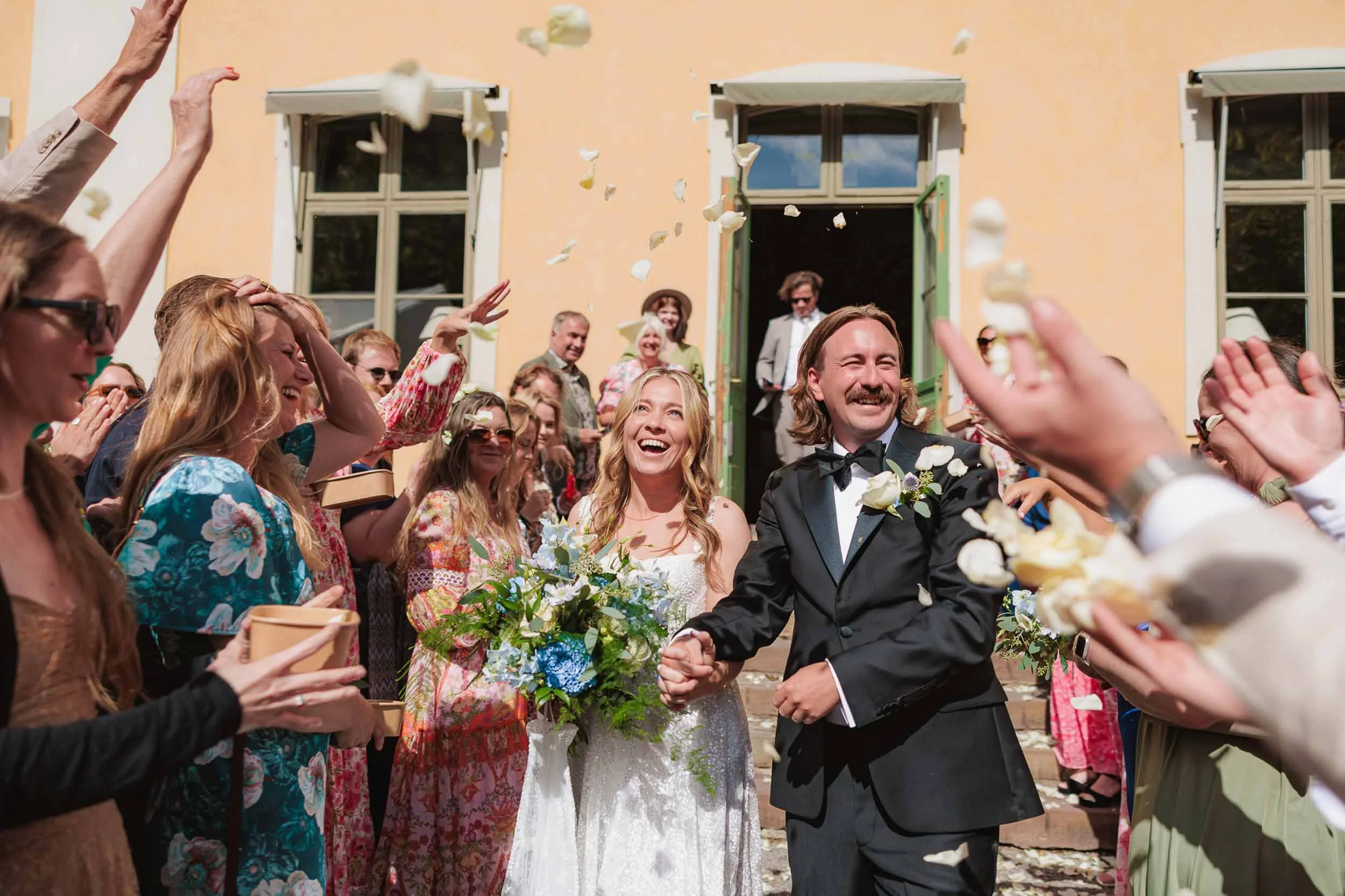 En nygiften par håller hand och skrattar bland gäster som kastar ris utefter en varmbelyst gul vägg med gröna fönsterluckor.