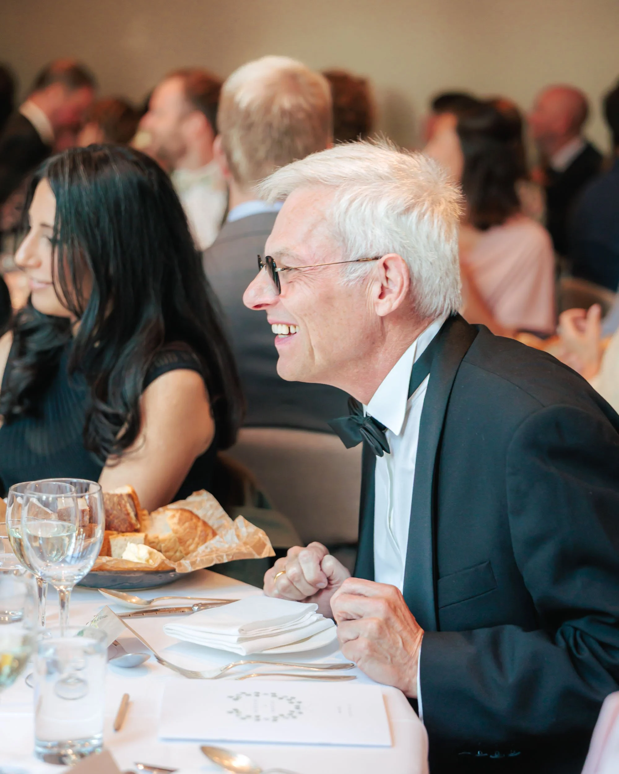 En glad äldre man i svart smoking med fluga, sitter vid ett bord på en formell sammankomst, med andra personer i bakgrunden.