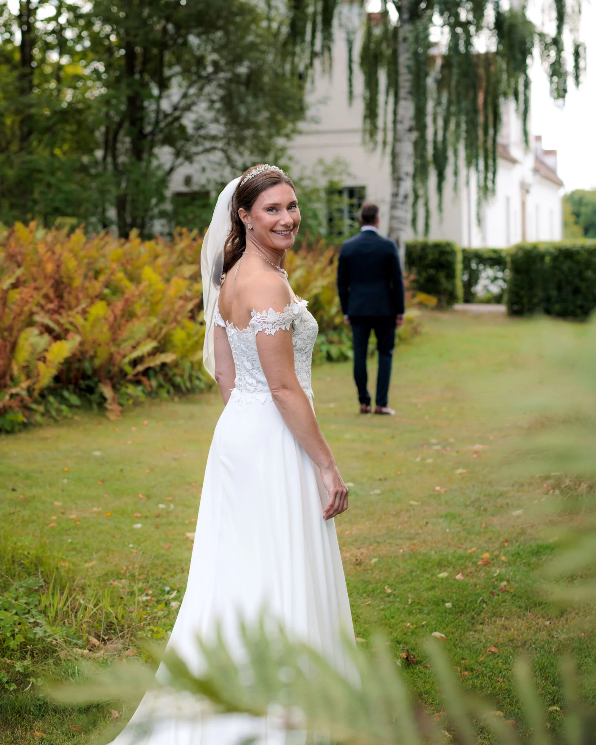 En glad brud i vit brudklänning och med slöja, står i en trädgård och vänder sig mot kameran, med en man i mörk kostym i bakgrunden.