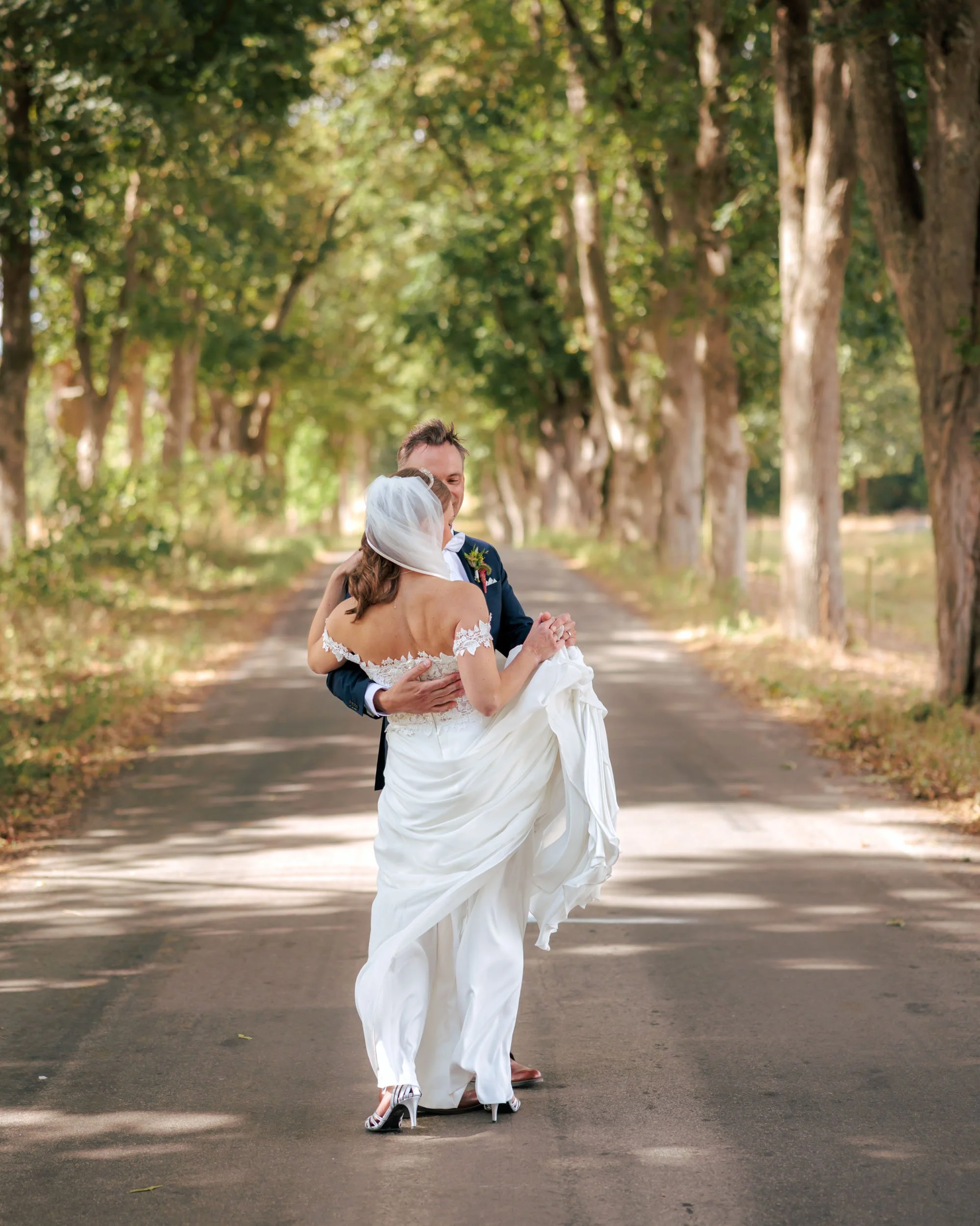 En nygiftemål på en landsväg omgiven av träd, där en man bär en mörk kostym och en kvinna i en vit bröllopsdräkt blir dansande.