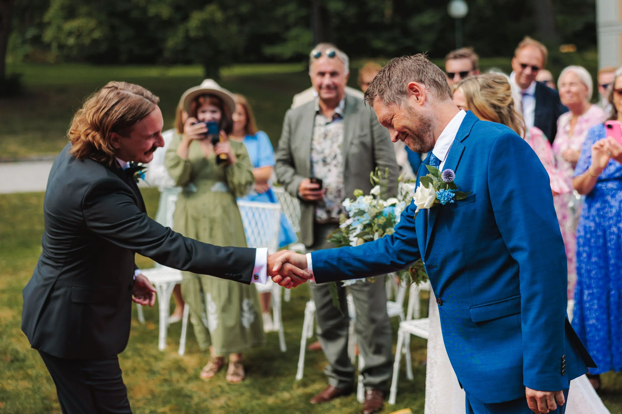 Två män i formella kläder skakar hand vid ett utomhusbröllop medan en grupp av glada gäster tittar på.