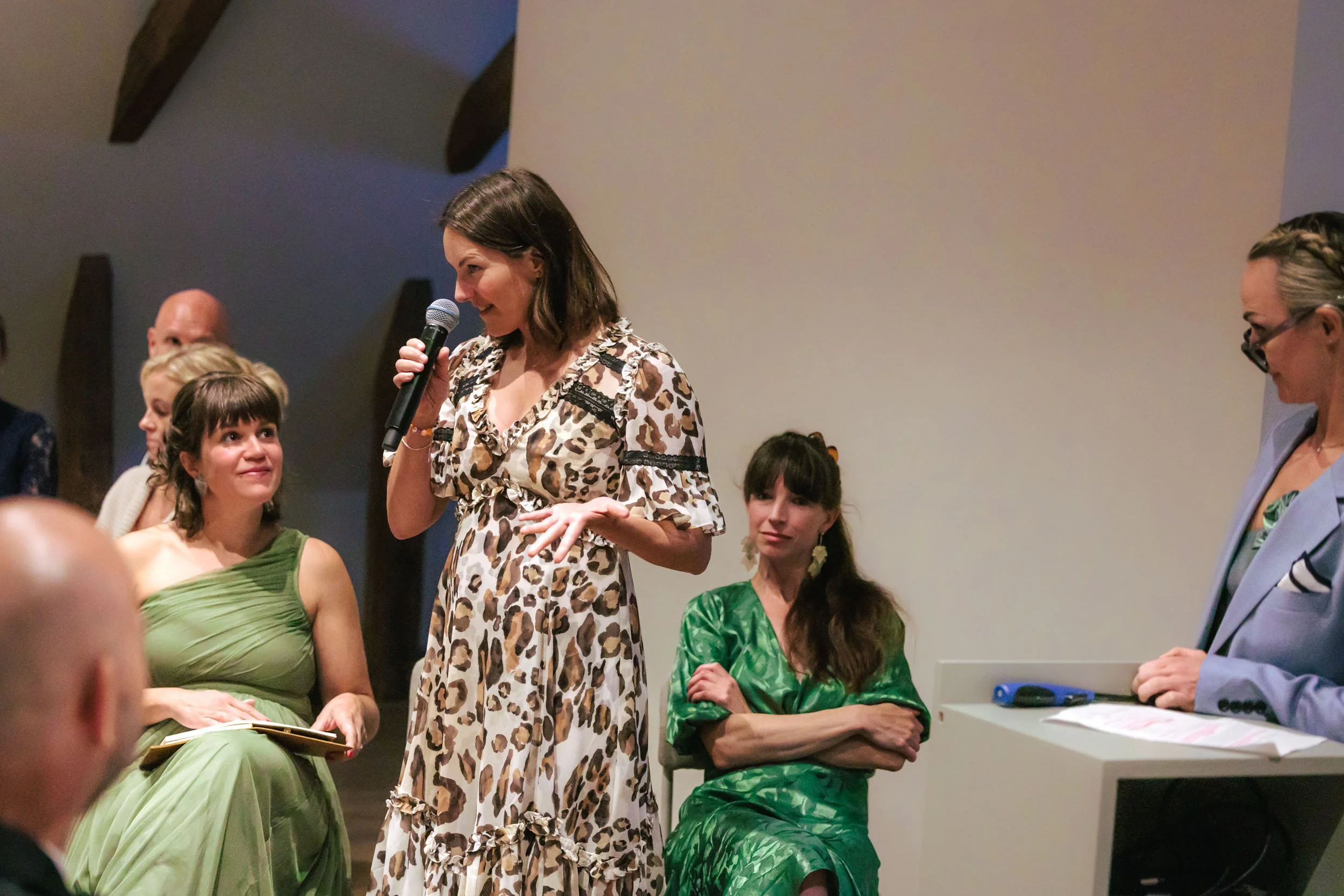 En kvinna i blommig klänning håller mikrofon och talar, omgiven av flera kvinnor som lyssnar.