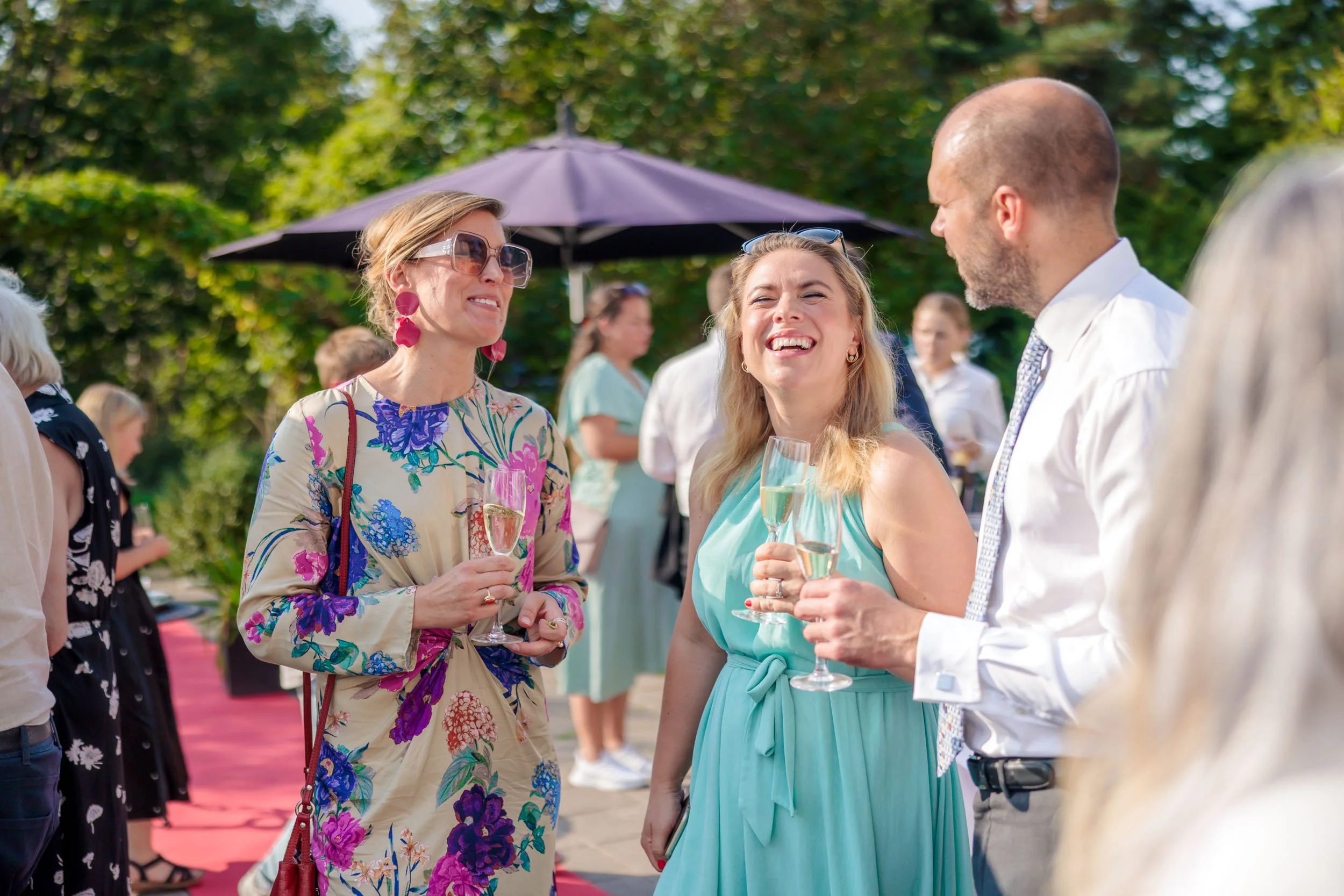 Tre personer som samtalar och skrattar på en utomhusfest, alla håller glas med champagne, i bakgrunden finns andra gäster och ett parasoll.