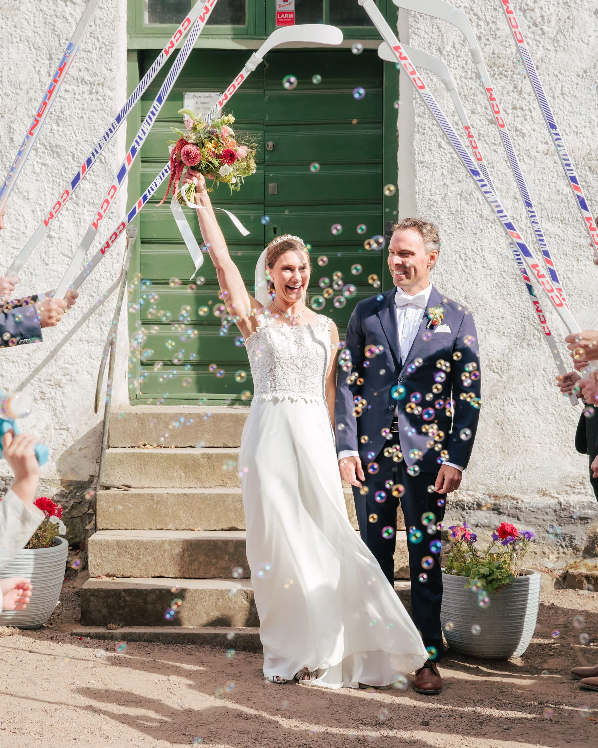 En brud och brudgum som firar giftermål utomhus framför en grön port, omringade av personer som skickar konfetti och bubblor, med blomkrukor på trappstegen.