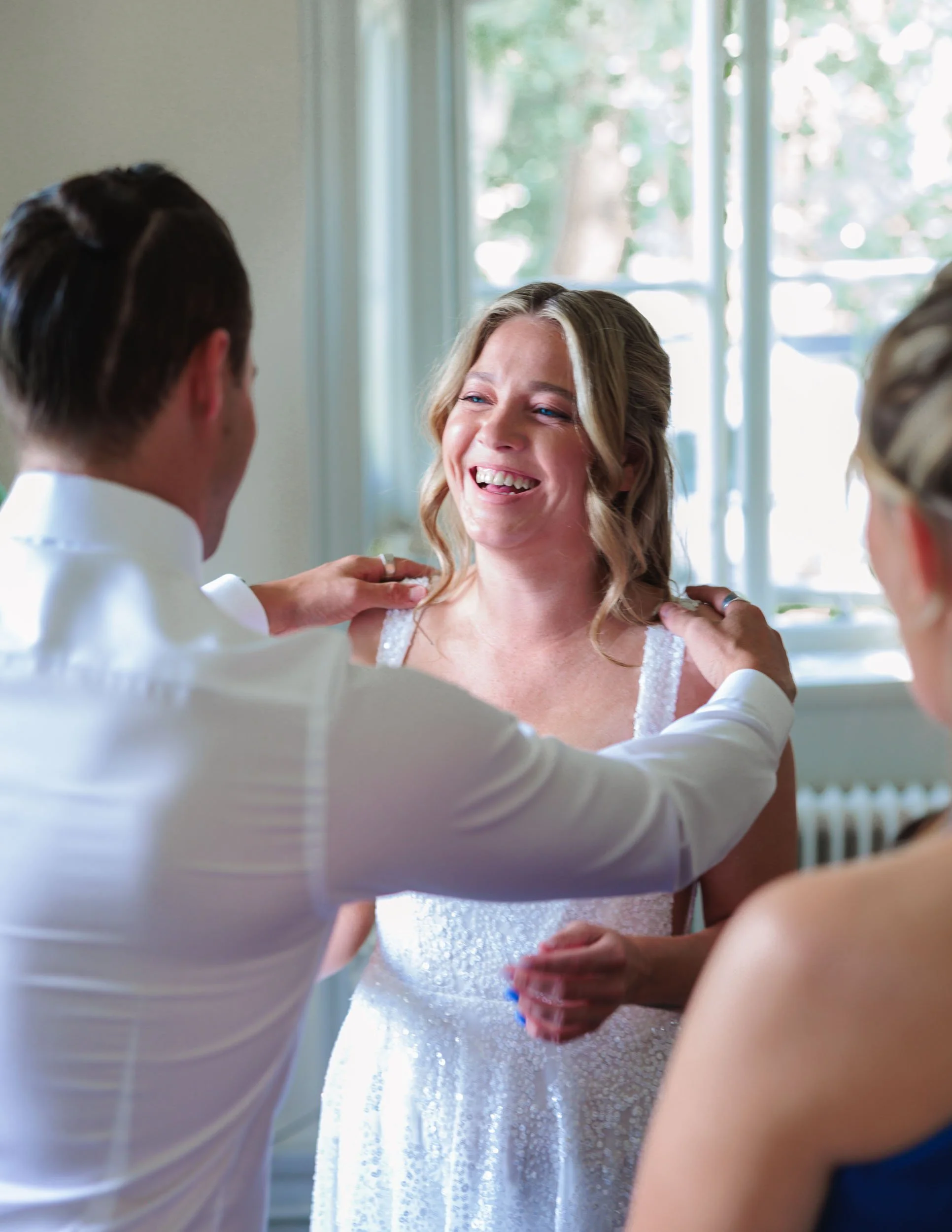 En kvinna i en bröllopsklänning ler medan två personer hjälper henne att justera klänningen.