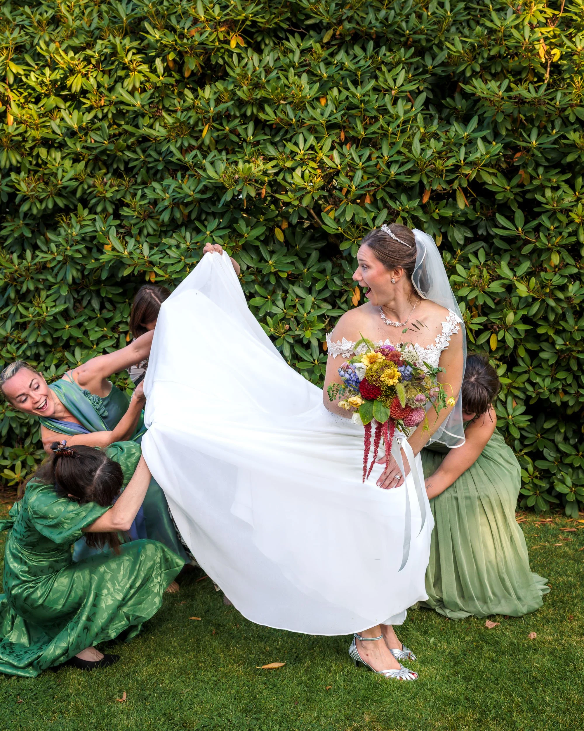 Brudgum och tre brudtärnor hjälper till att lyfta brudens klänning utomhus, med grön buske i bakgrunden. Bruden bär en vit klänning och håller en färgglad blomsterbukett.