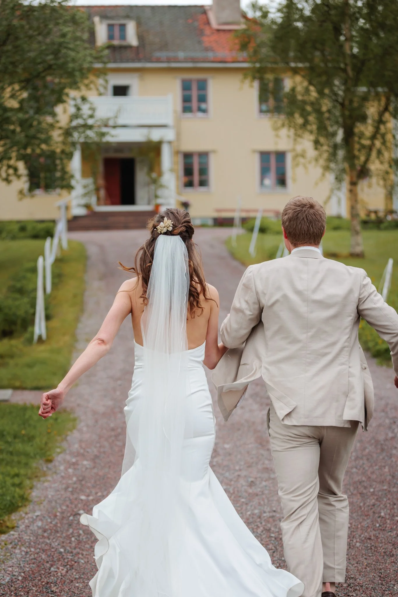 Brud och brudgum springer hand i hand uppför gruspiren mot gul gårdsbyggnad på molnig bröllopsdag