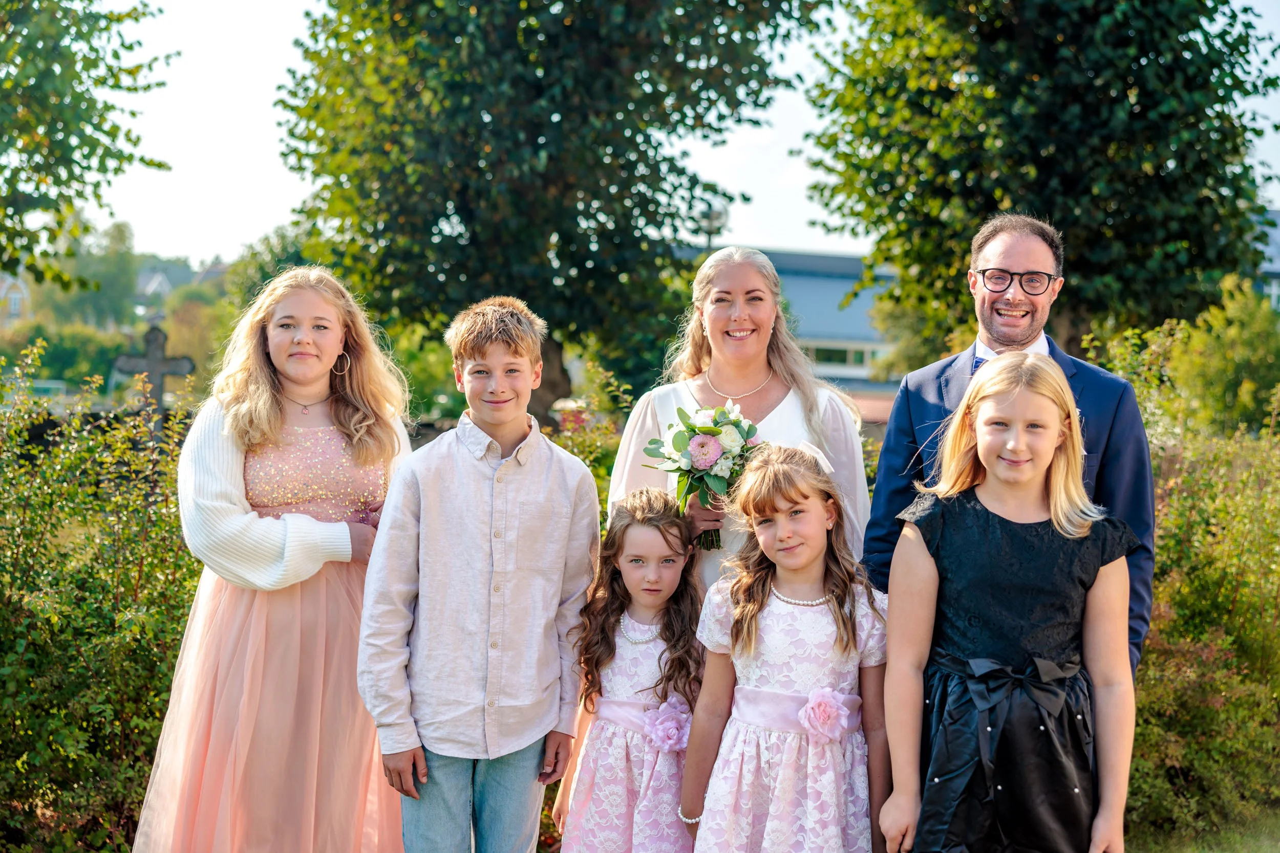 En familj som står tillsammans utomhus, troligen under en fest eller ceremoni, med träd och hus i bakgrunden.