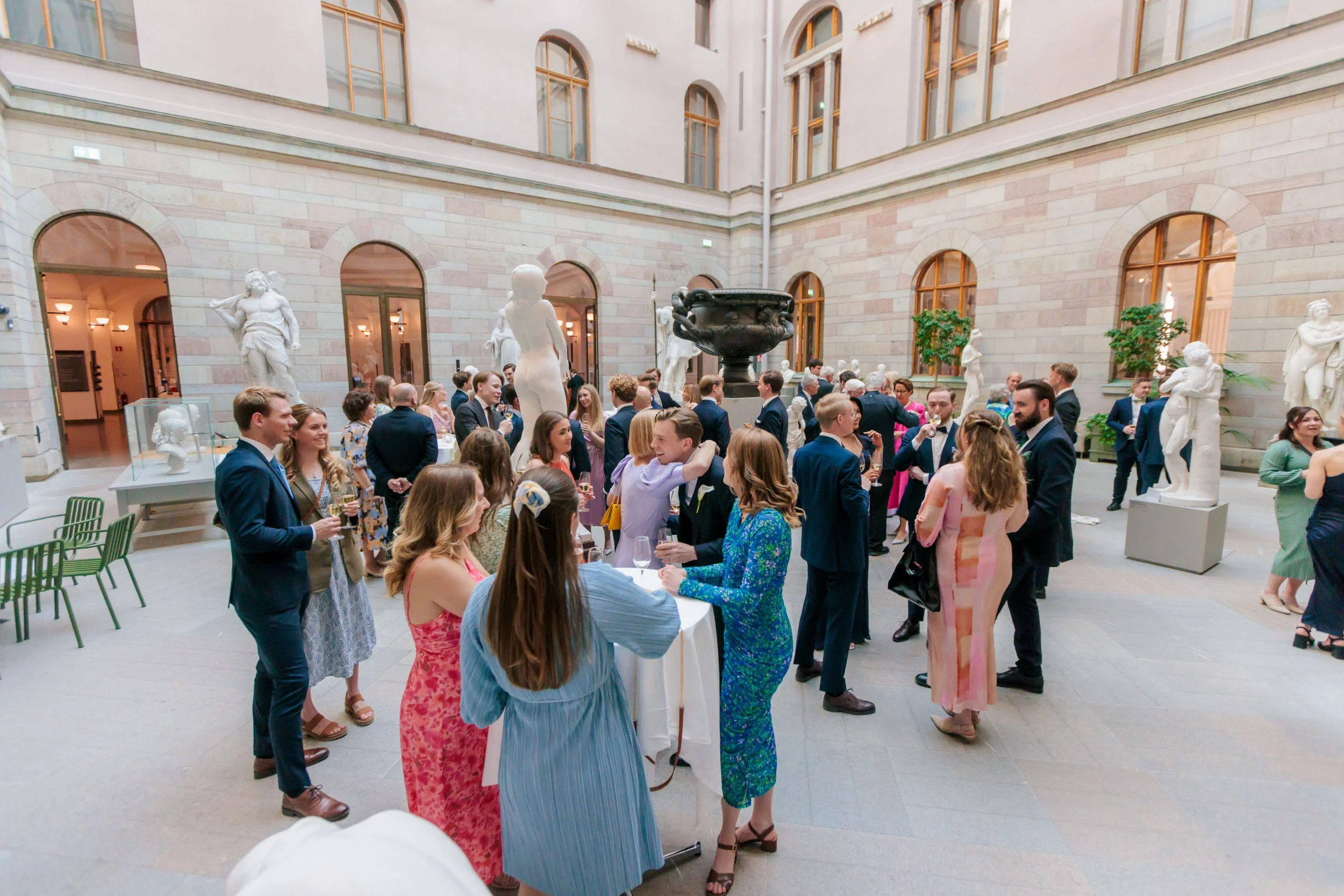 Bröllopsgäster som minglar i en stor ljus sal med statyer; information om halvdagspaket för bröllopsfilmning i 6 timmar.