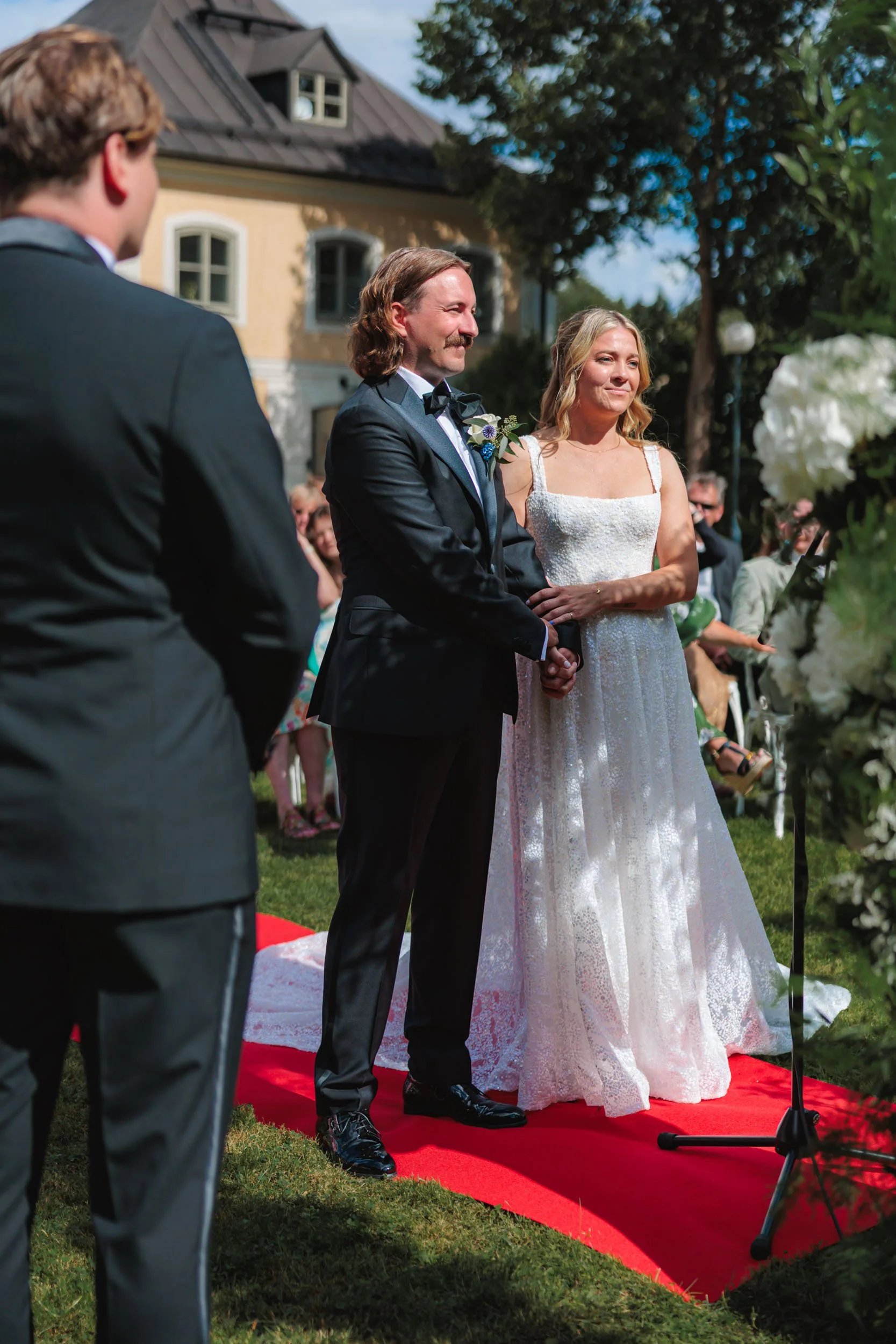 Brudparet står på en röd matta under ett utomhus bröllop, med ett par personer i förgrunden, där mannen i svart tuxedo och kvinnan i vit brudklänning håller handen och ser glada ut.