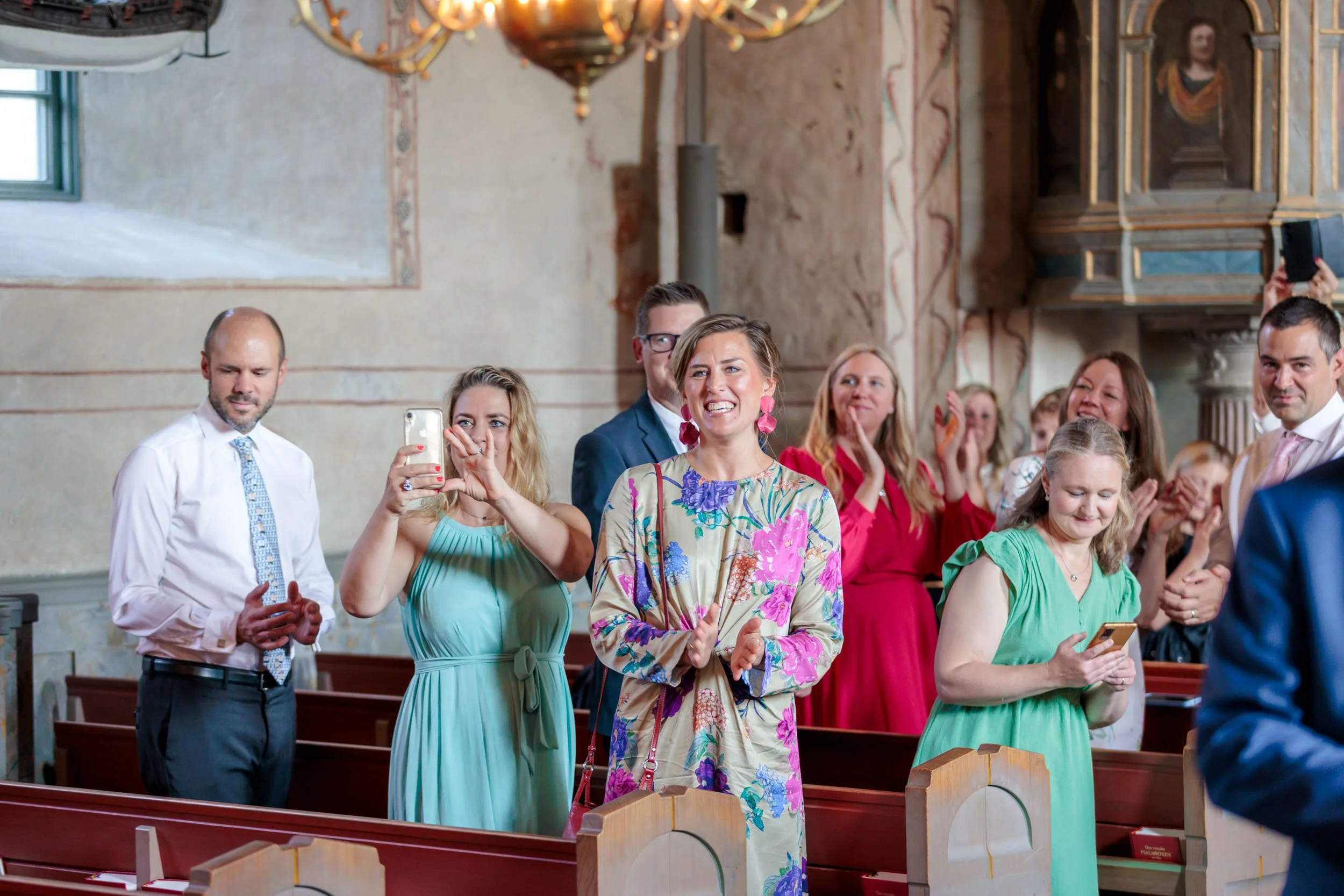 En grupp människor, troligen i en kyrka, bekvämt klädda, som verkar delta i ett bröllops- eller ceremoniförrättande, med många som tar bilder.