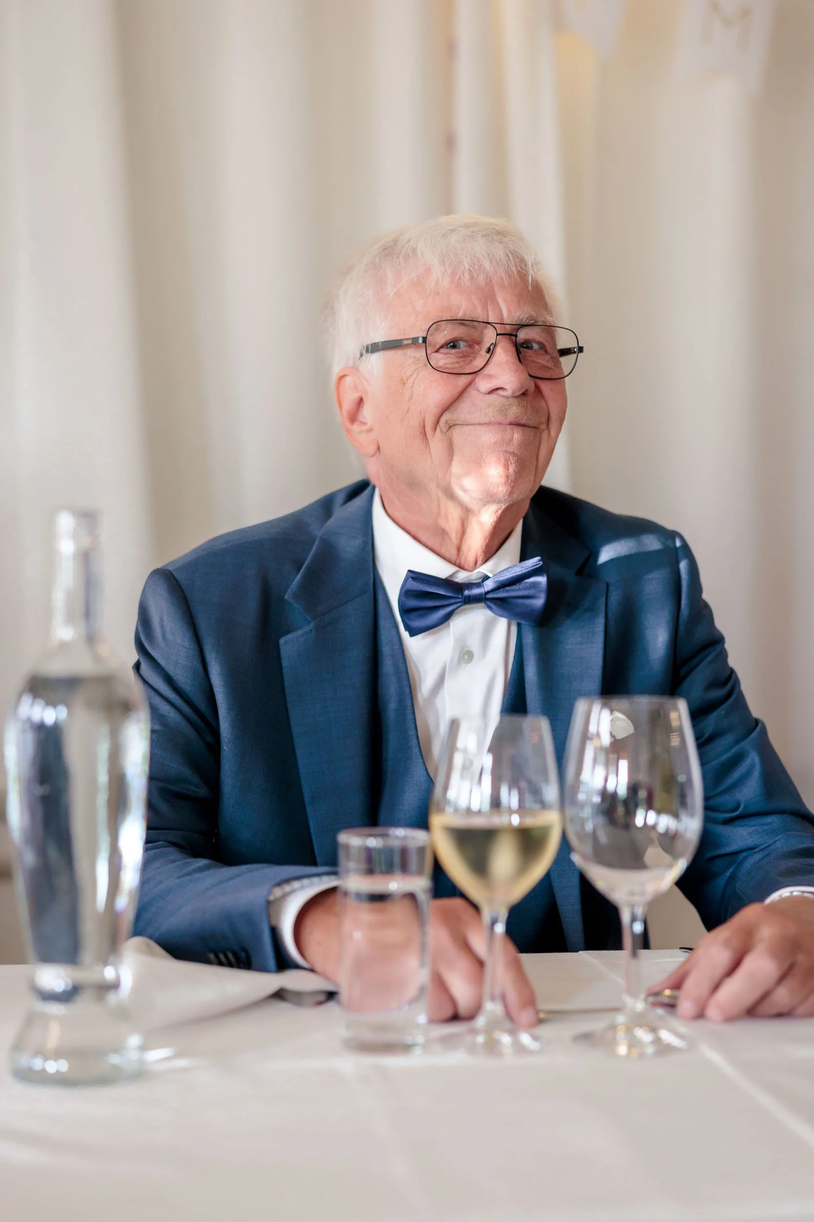 En äldre man i kostym och fluga sitter vid ett bord med olika glas vin och vatten, ler och tittar framåt.