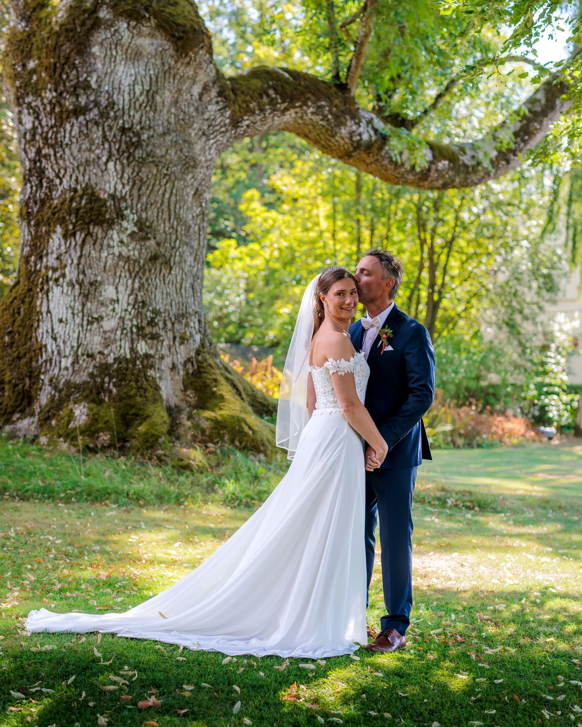 En brud och brudgummen står tillsammans under ett stort träd i en trädgård, omgivna av gröna löv och sollys.