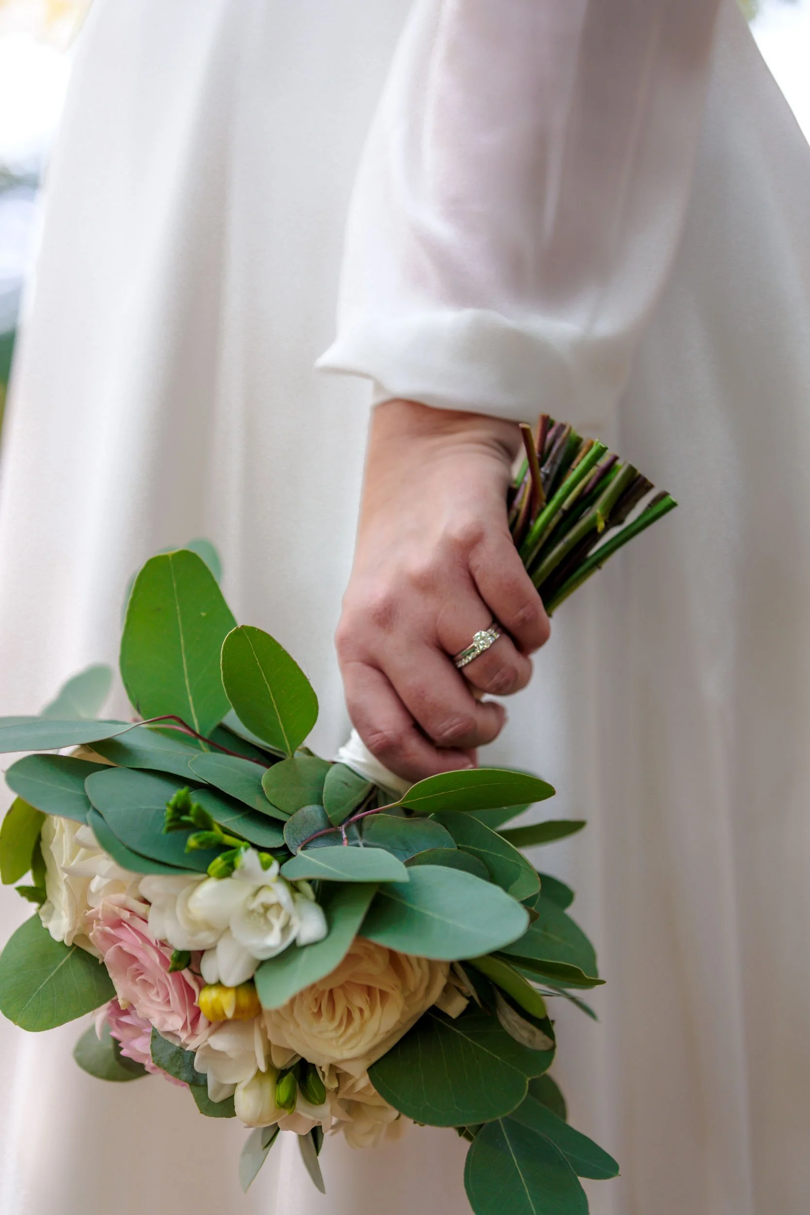 En hand med vigselring håller ett brudbukett med gröna blad och ljusa blommor, klädd i ett vitt, långärmat plagg.