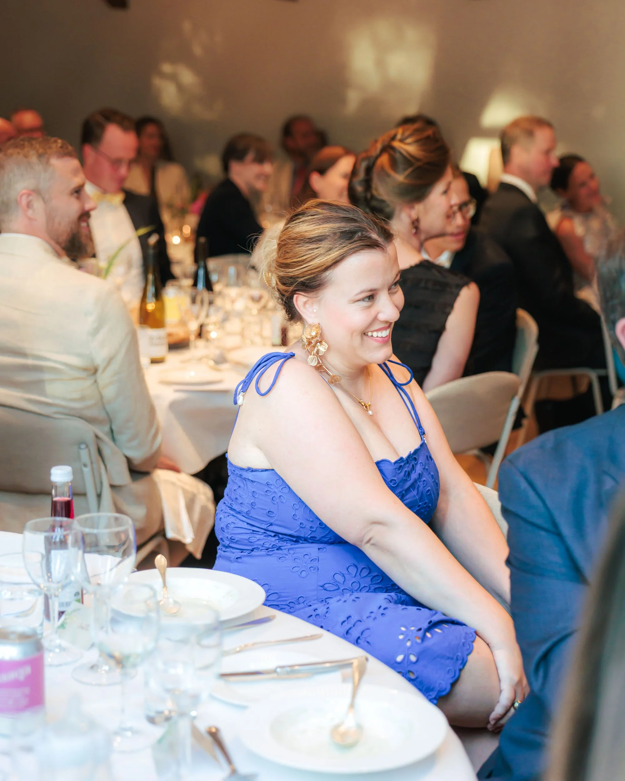 Människor sitter vid ett bord under en fest, kvinnor och män i formella kläder, inklusive en kvinna i en blå festklänning som skrattar.