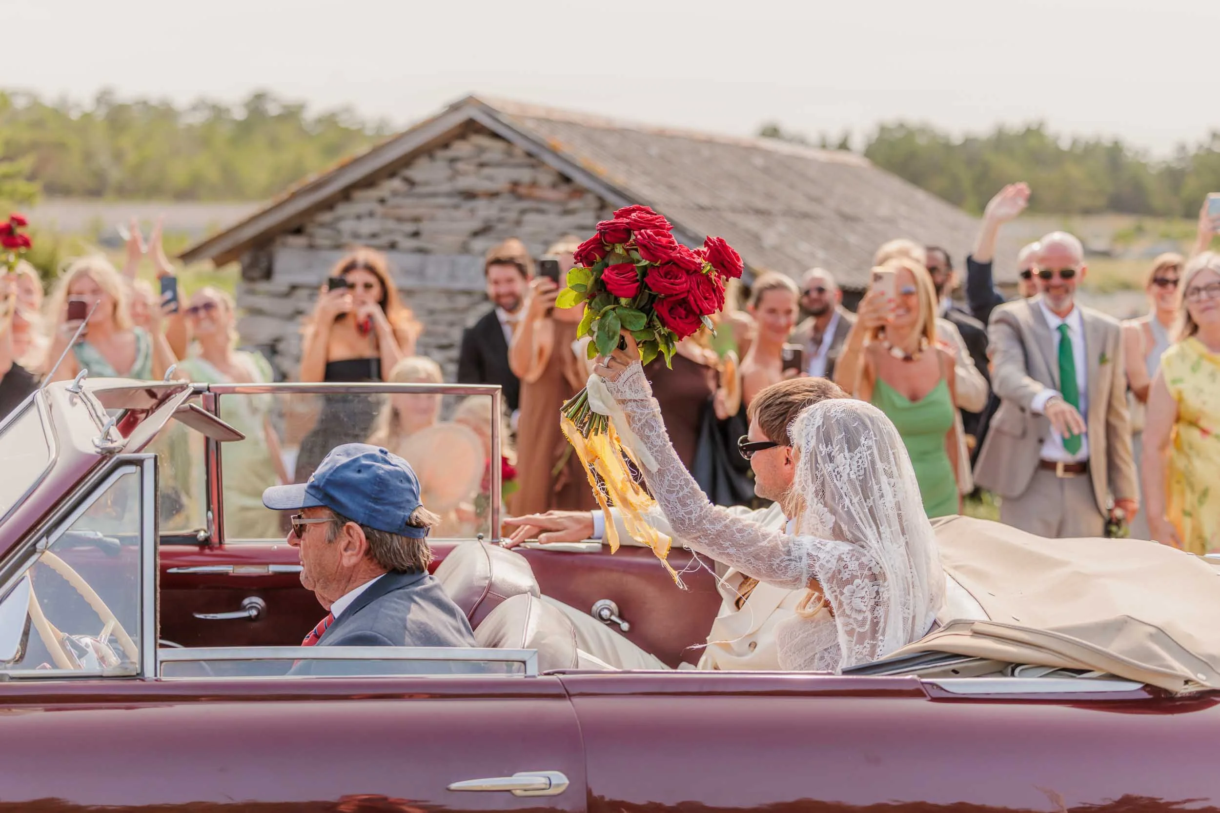 Brud vinkar från öppen bil medan gäster fotograferar under bröllopsfirande utomhus