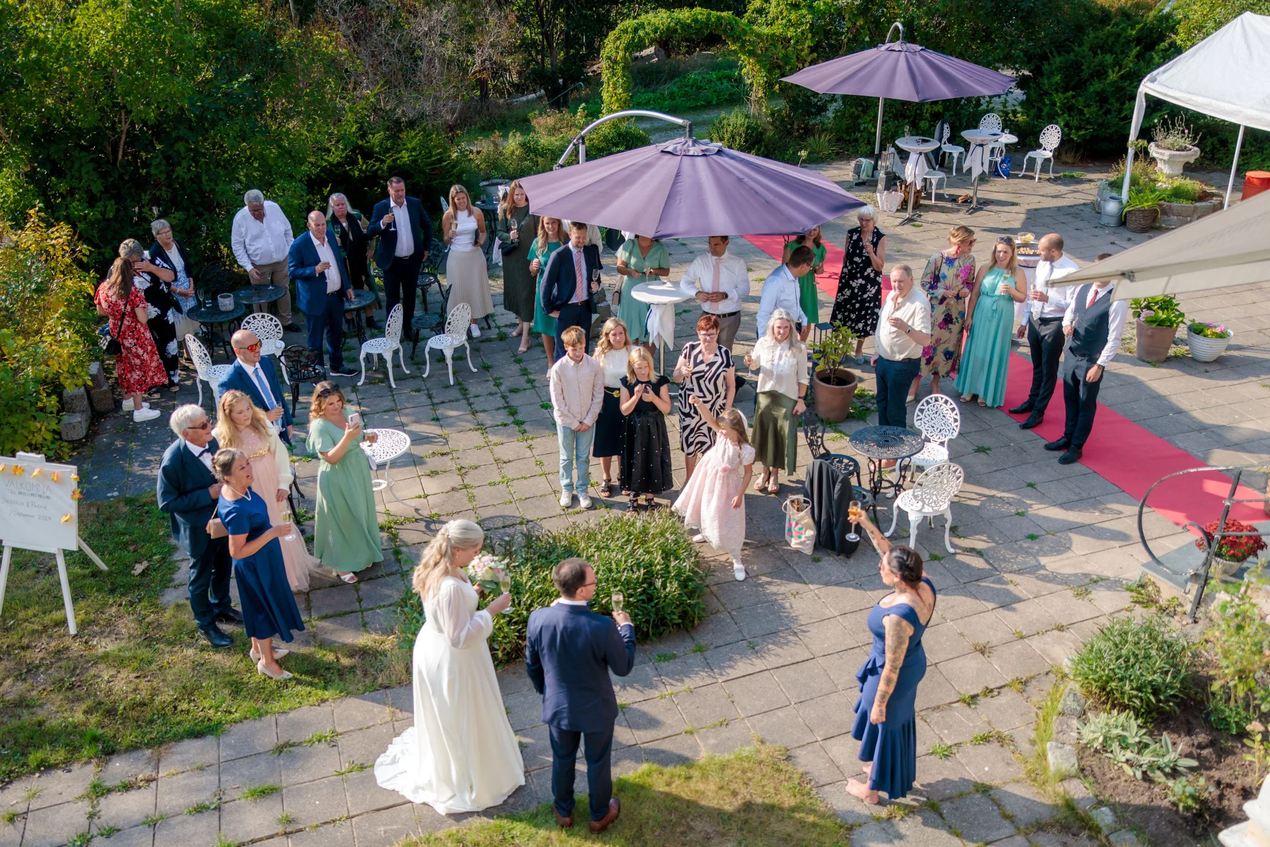Gäster vid ett utomhusbröllop samlas på en stenlagd terrass med gröna buskar och träd att växa runt omkring. Den ängsliga ceremonin pågår i förgrunden framför ett brudpar, där en kvinna i en vit brudklänning och en man i kostym håller tal. Gäster bär