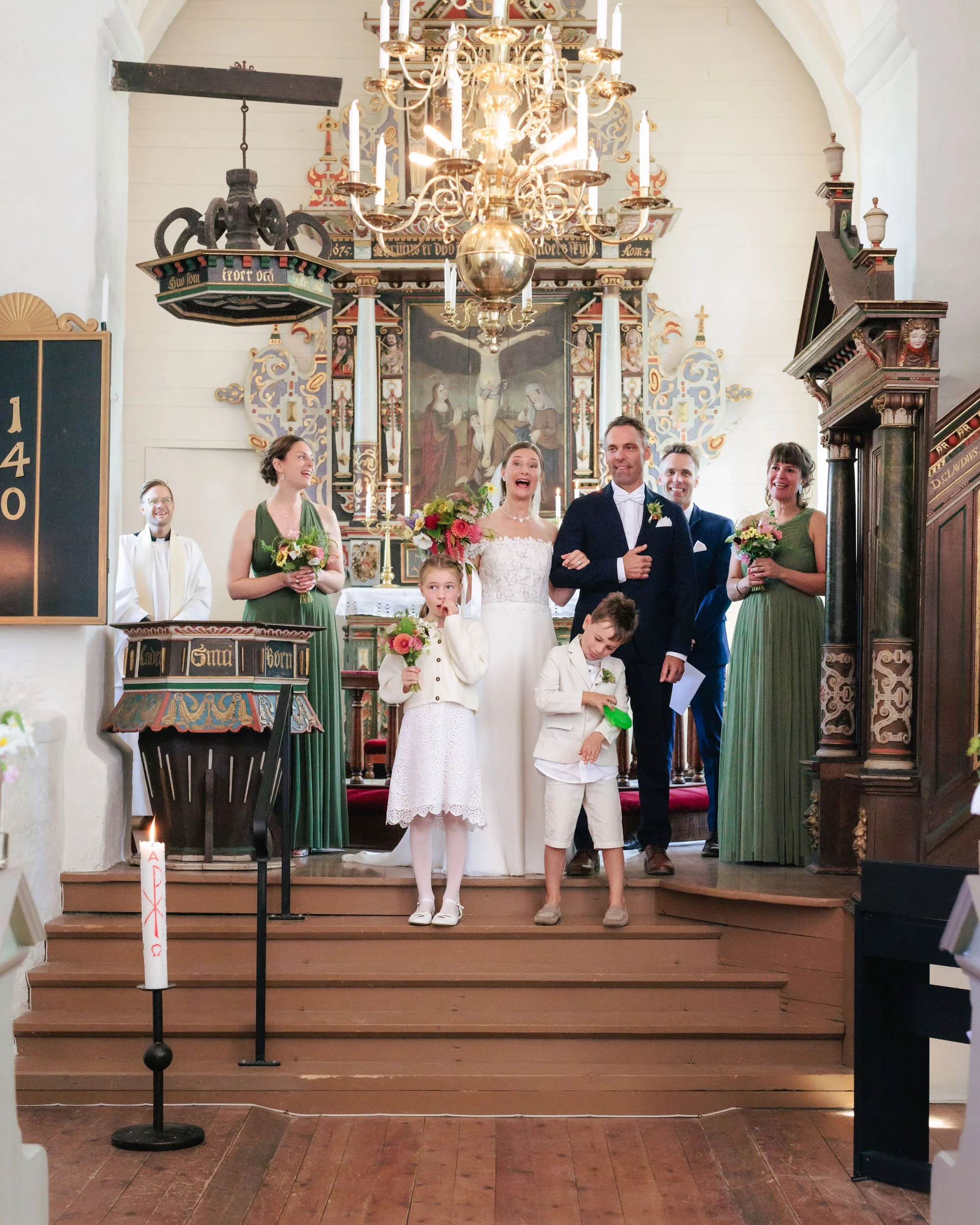 En brud och en brudgömne står tillsammans på en kyrklig altarbänk, omgivna av tre kvinnor och två barn, vid ett bröllop. Alla är glada, och bakgrunden är en kyrka med ljusblå väggar, älvgolv och ett stort silverkronljus.