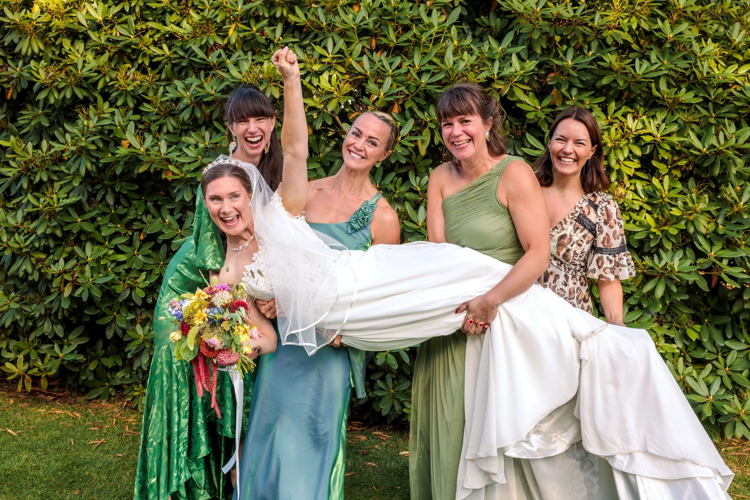 Fem kvinnor står mot en buske, en av kvinnorna ligger i luften och hålls av de andra. Den kvinnan i mitten bär en brudklänning och håller en bukett blommor. Alla är glada och skrattar.