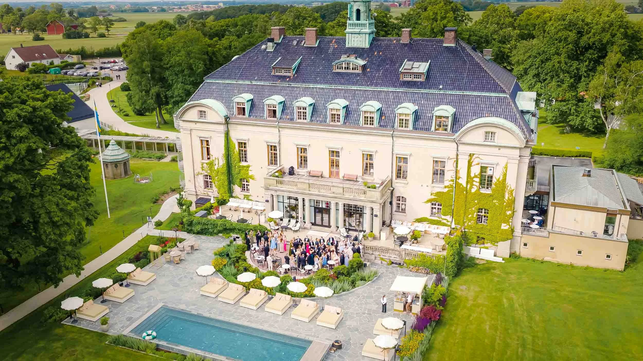Drönarfoto över ett pampigt vitt slott med bröllopsfest på gården och poolområde.
