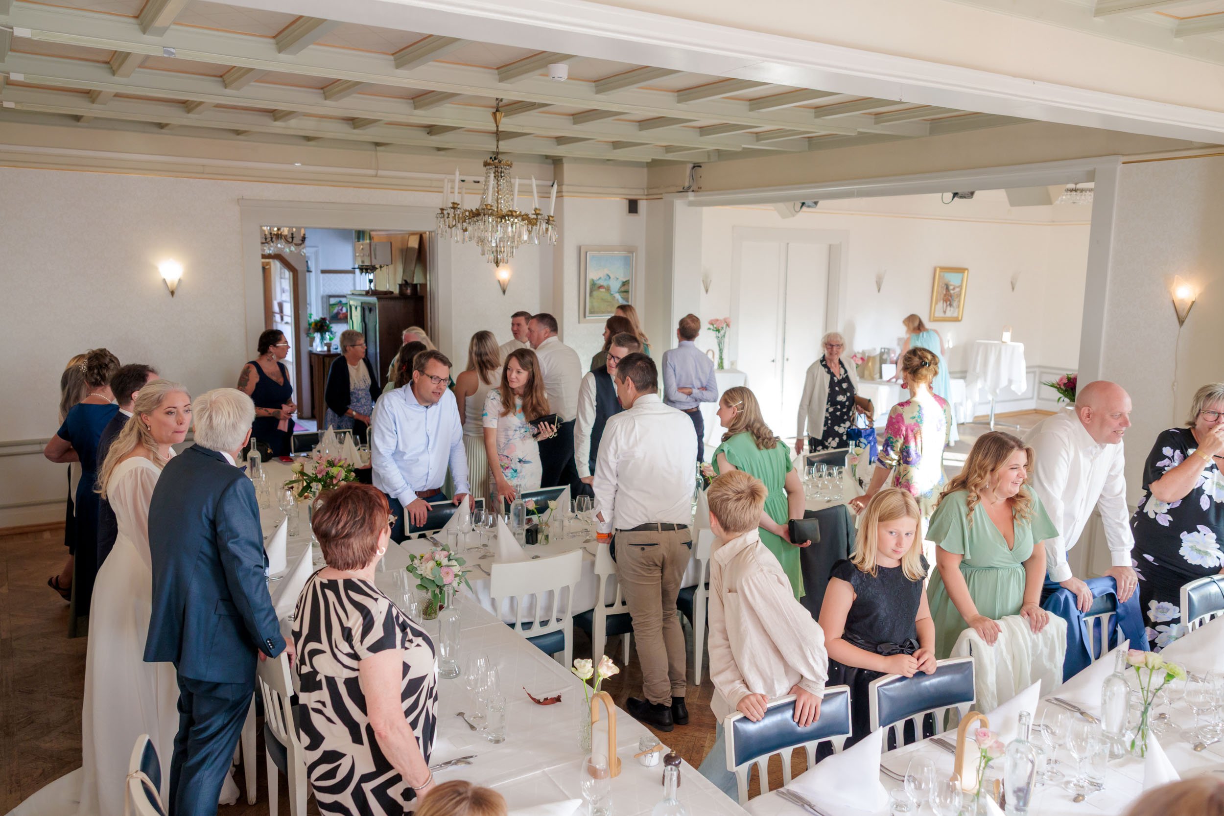 Många personer samlas i ett festligt rum för ett evenemang, troligen ett bröllop, med dukade bord, blommor och ljus.