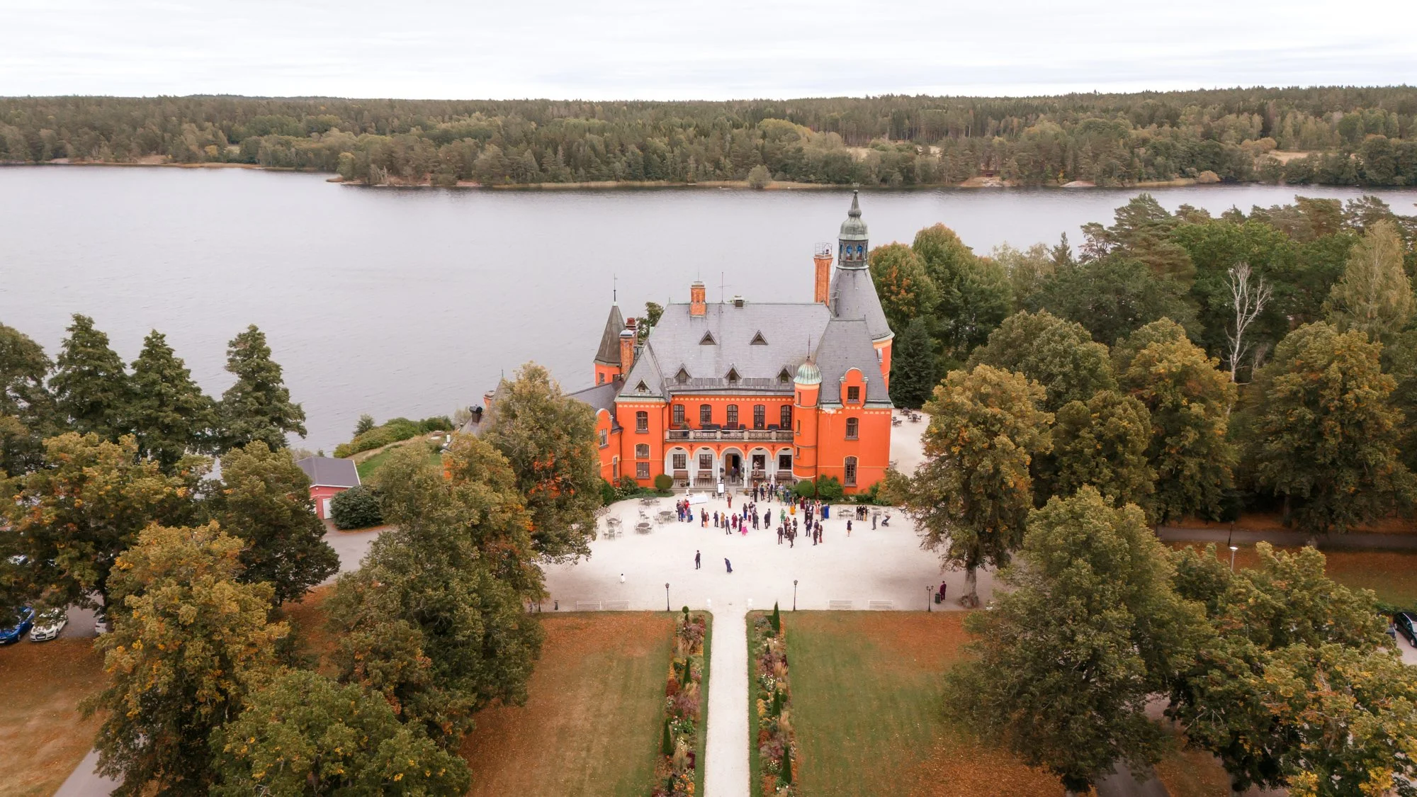 Drönarvy över rött slott vid sjö med bröllopsgäster på gårdsplan – bröllopslokal slott