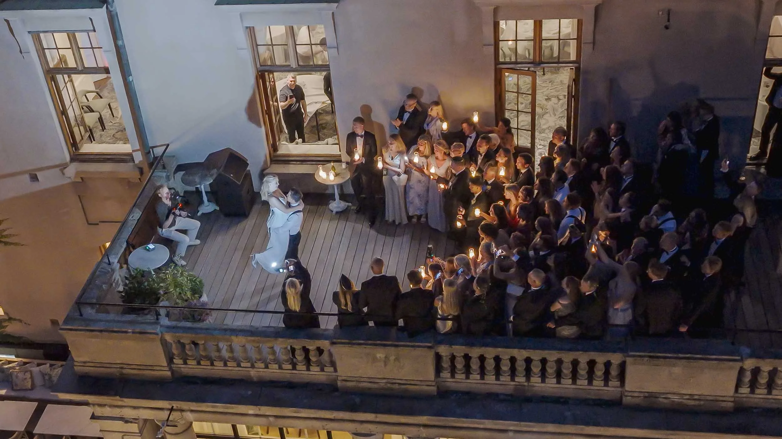 A group of people gathered on a balcony in the evening sun; many are holding candles, while a man and a woman dance in the center. An outdoor setting with open windows, and people dressed for the evening.