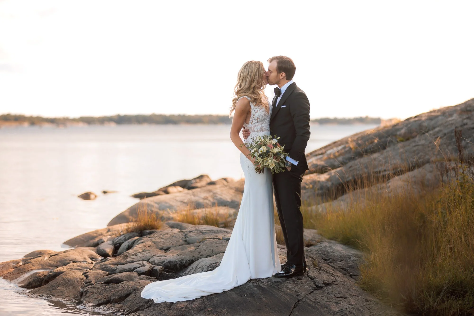 Brudpar som kysser varandra på klippor vid vattnet i det mjuka kvällsljuset.
