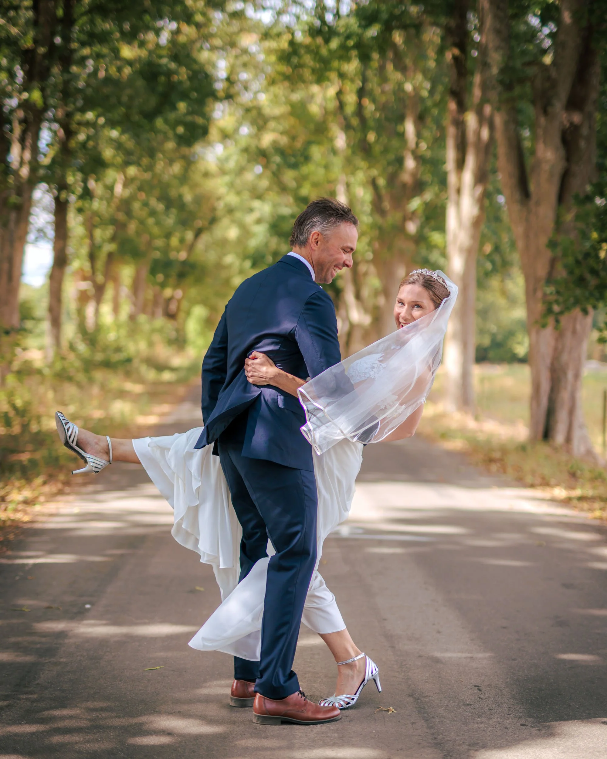 En brud och en brudgöm hjælps åt i en skogsstig. Brudgummen bär bruden i sina armar. Bruden ler mot kameran, och båda är klädda i bröllopsutstyrsel.