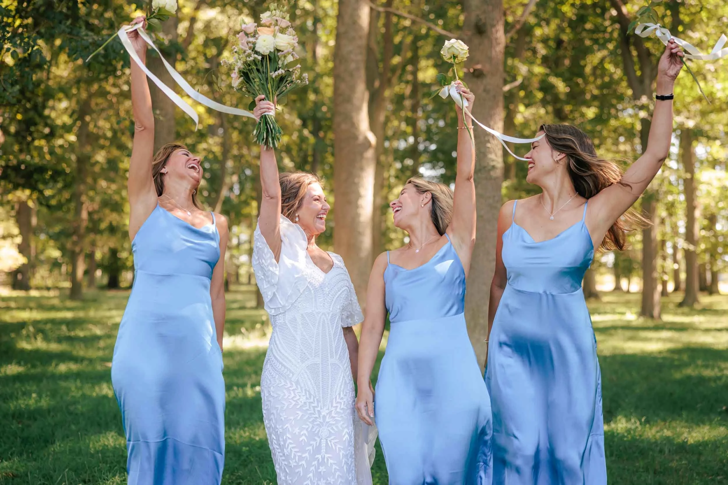 Fyra kvinnor i blå klänningar och en kvinna i en vit klänning står tillsammans i en park, firar med blomsterbuketter och snöror, under en solig dag.