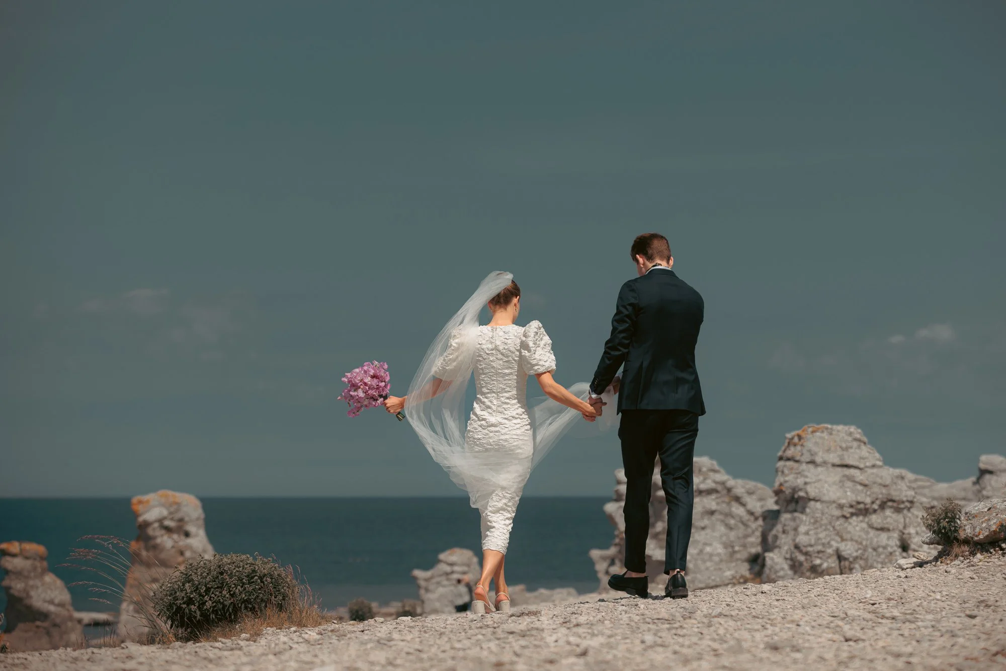 Brudpar håller hand och går mot havet bland raukar på Gotland med slöja i vinden