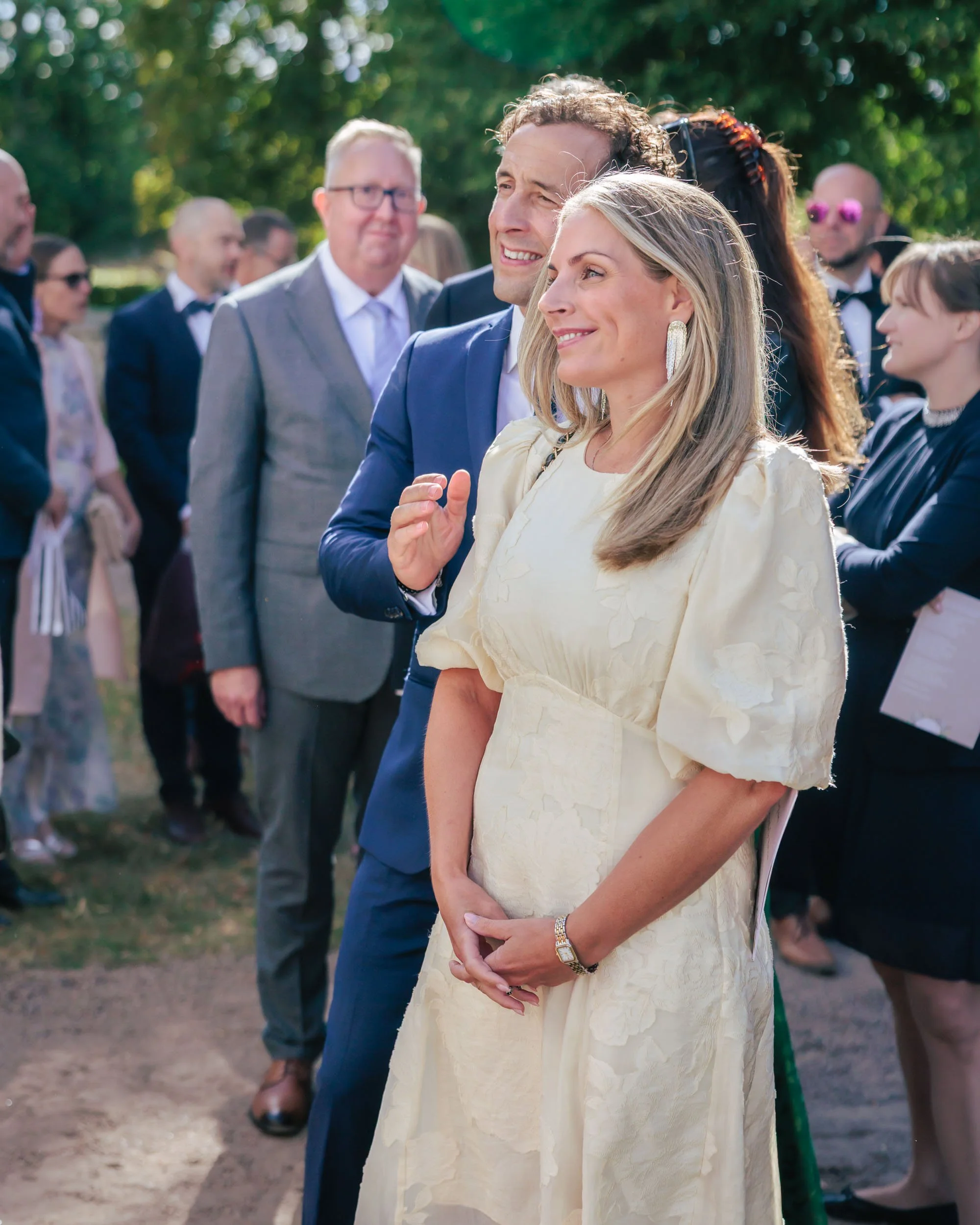 Grupp människor på ett utomhusevenemang, en kvinna i en ljusgul klänning och en man i blå kostym i förgrund.