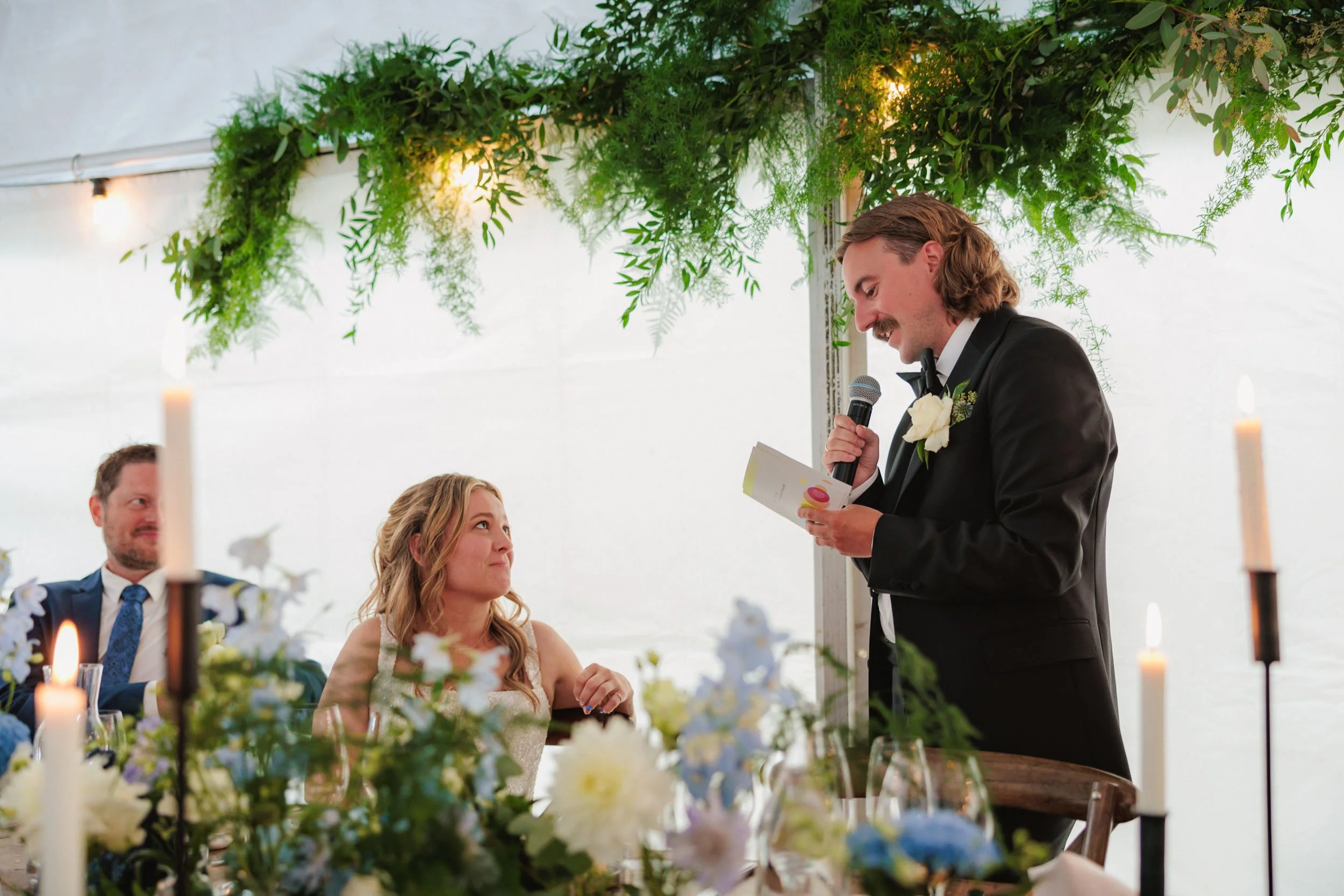 Brudgummen ger ett tal vid sitt bröllop, han håller ett mikrofon och ett papper, medan bruden och gäster lyssnar.