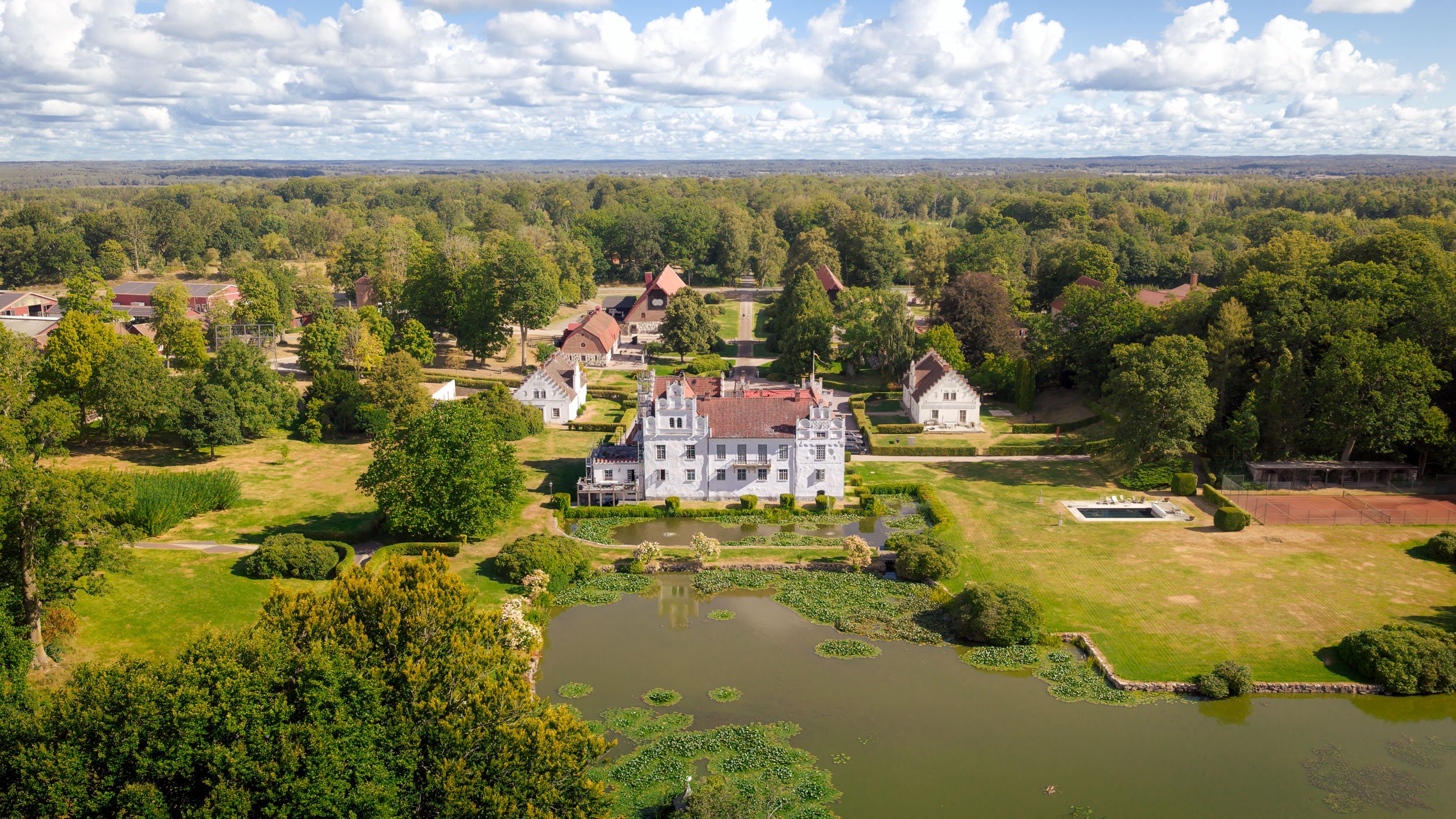 En flygbild av en stor herrgård med en trädgård, vatten och omgivande skog, under en molnig himmel.