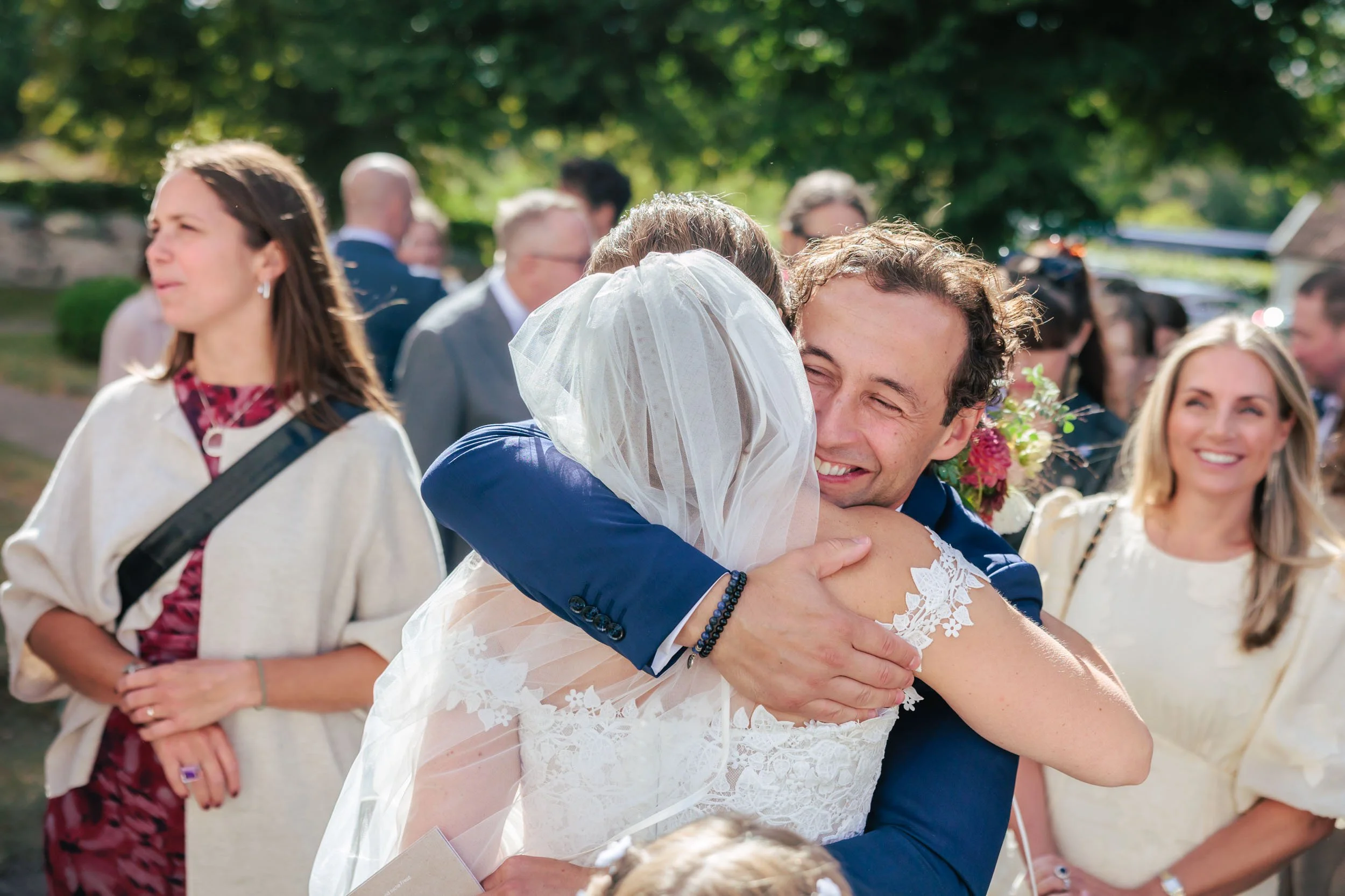 En man och en kvinna kramas lyckligt, på ett utomhusbröllop, med flera gäster i bakgrunden.