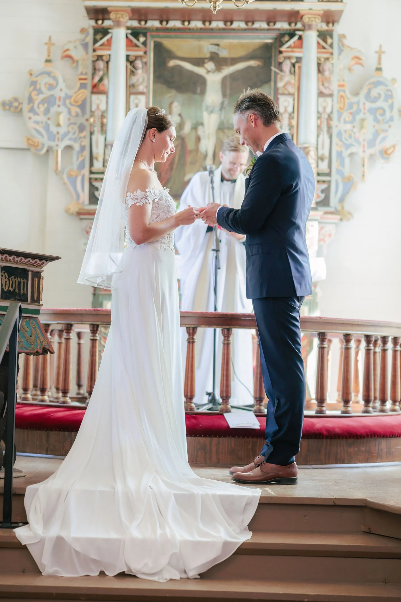 En brud och en brudgum som står på en altare under ett bröllopsnatt, de håller händer och ser ner på varandra, med en präst i bakgrunden i en kyrka.