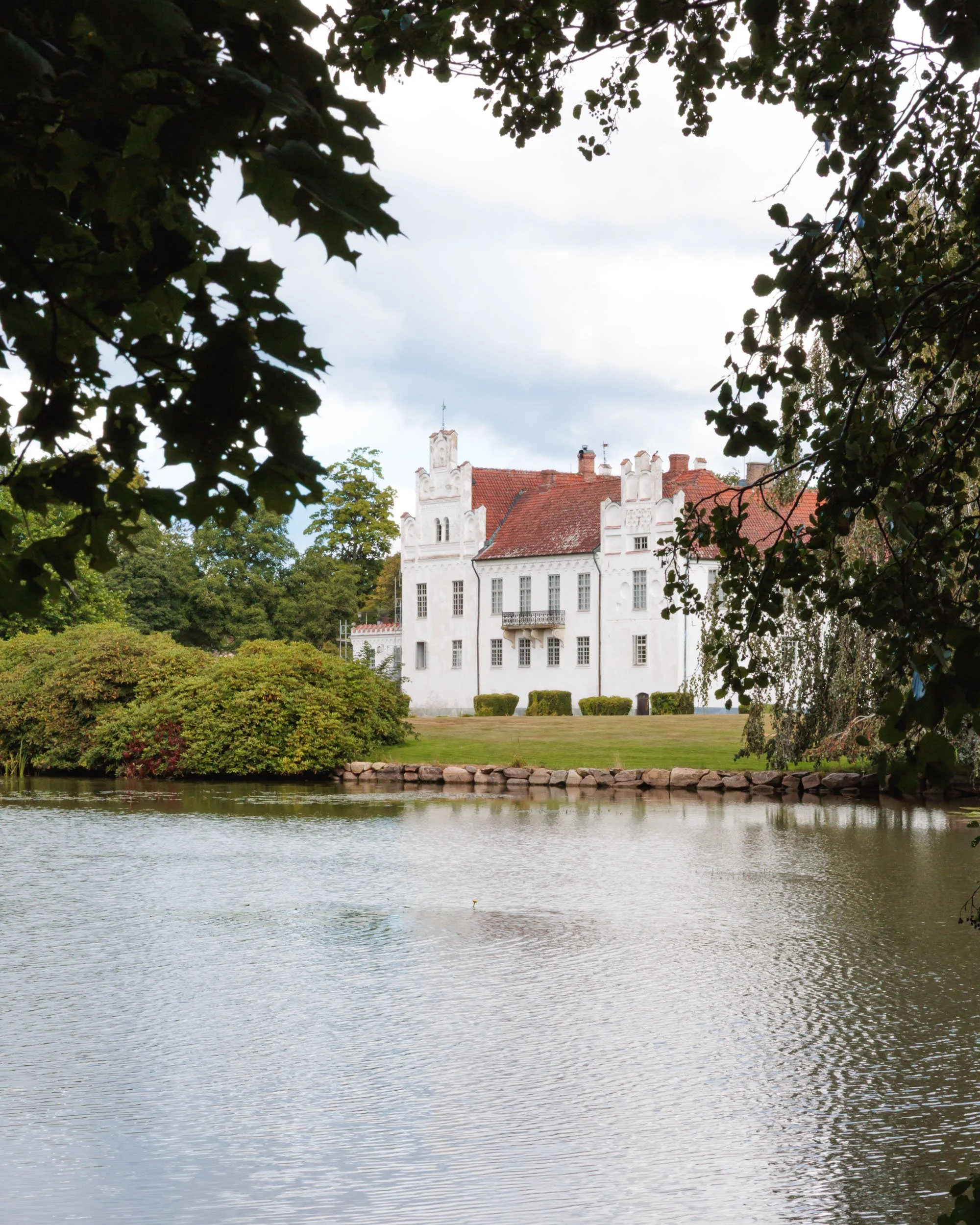 En vit herrgård med röda tak som ligger vid en sjö, omgiven av träd och buskar, skymd delvis av grönskande löv i förgrunden.