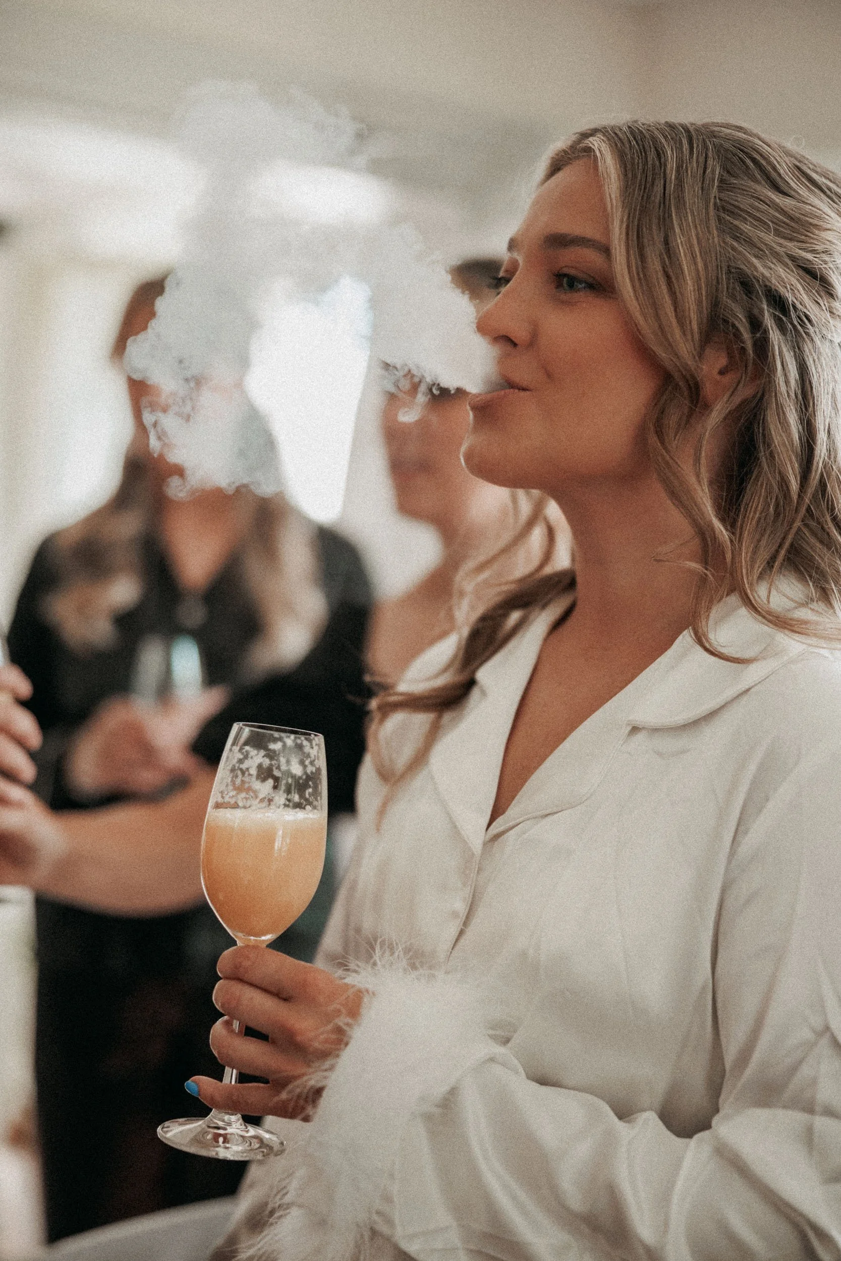 En kvinna med blont, lockigt hår som håller ett glas mousserande vin eller champagne, med rök eller ånga som stiger från glaset och henne, i en social sammanhang med andra personer i bakgrunden.