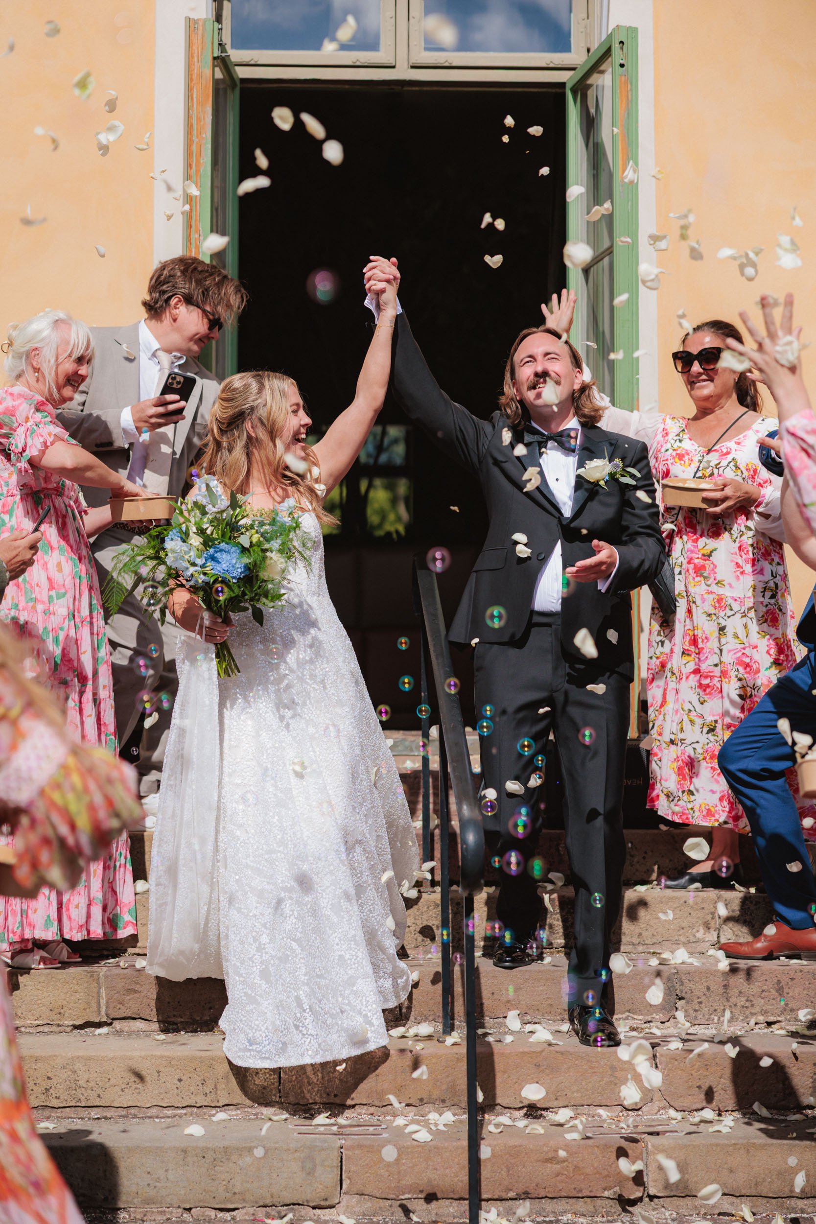 Ett brudpar som firar på trappan utanför en byggnad, omgivna av vänner och familj som kastar konfetti. Bruden håller en bukett blommor och väntar på att brudgummen, som i en svart tuxedo, ska höja sin hand i triumf.
