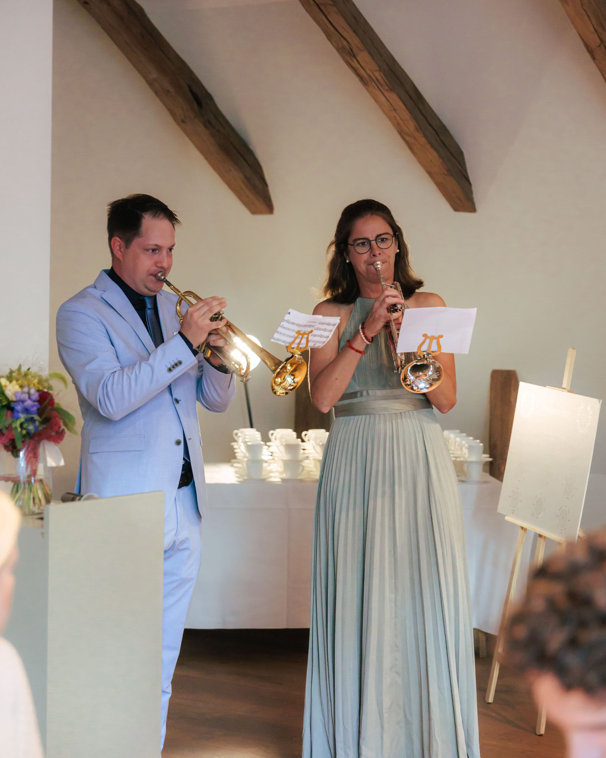 Två personer spelar trumpet under ett bröllop eller fest, en man och en kvinna, båda i formella kläder, i ett rum med träbjälkar i taket.