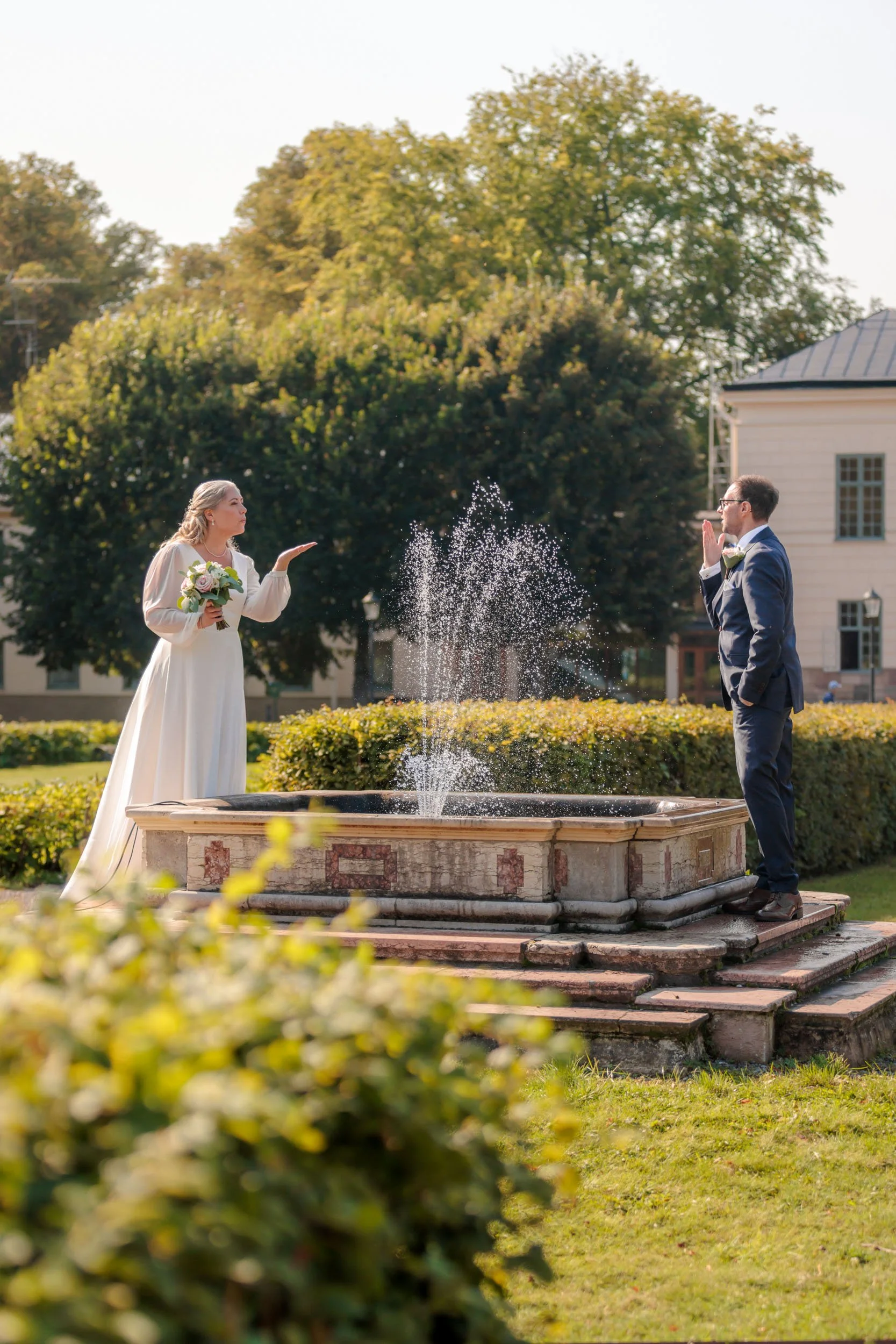En brud och en brudgum står mitt emot varandra i en trädgård, med en liten fontän emellan dem. Brudens hand är utsträckt som om hon kastar något mot brudgummen, som har båda händerna samman i en gest av omfamning. Solen lyser och det är gröna träd oc