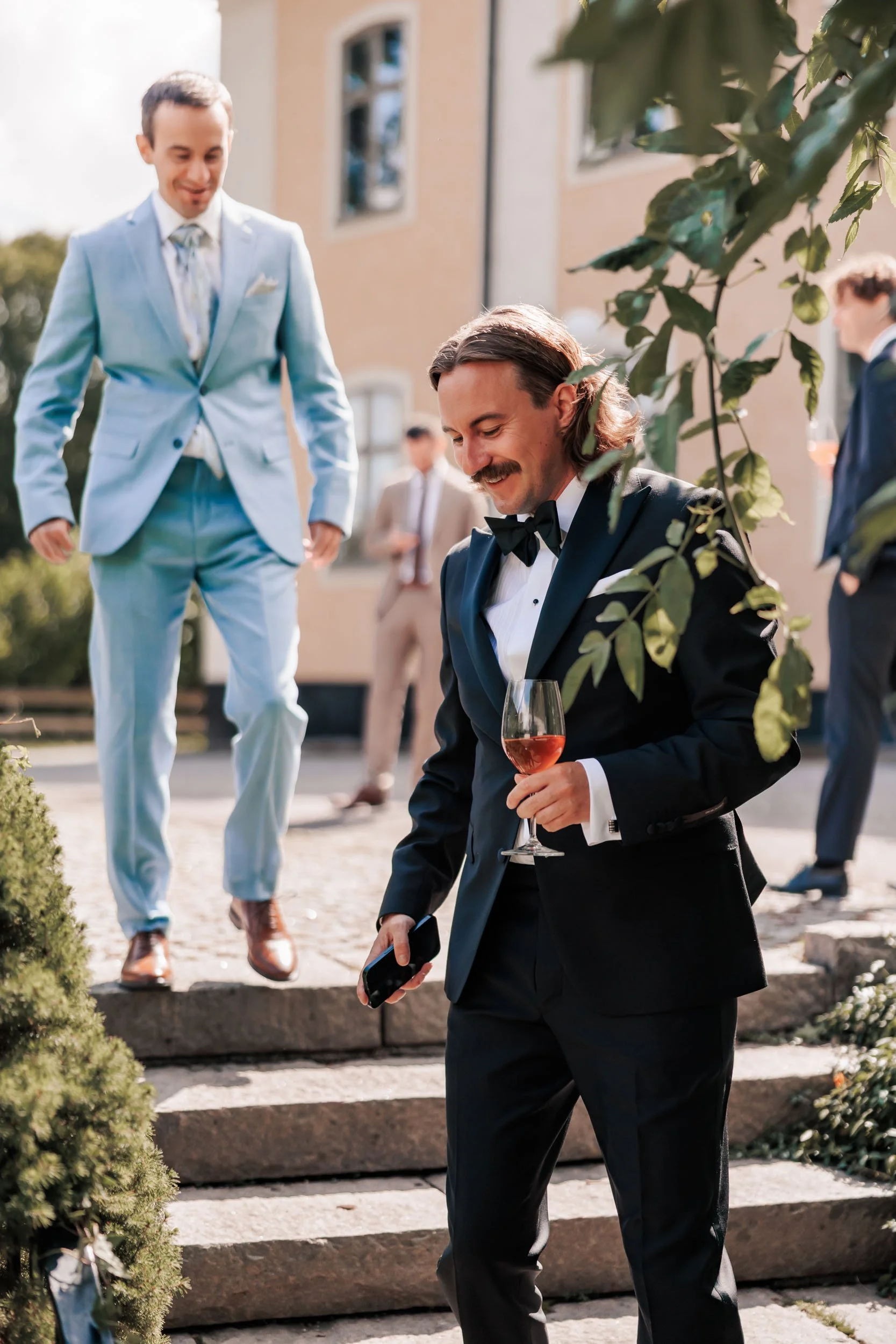 Tre män i formella kläder, en i svart smoking med glas rosévin, en i ljus blå kostym och en i beige kostym, utomhus vid en fest.