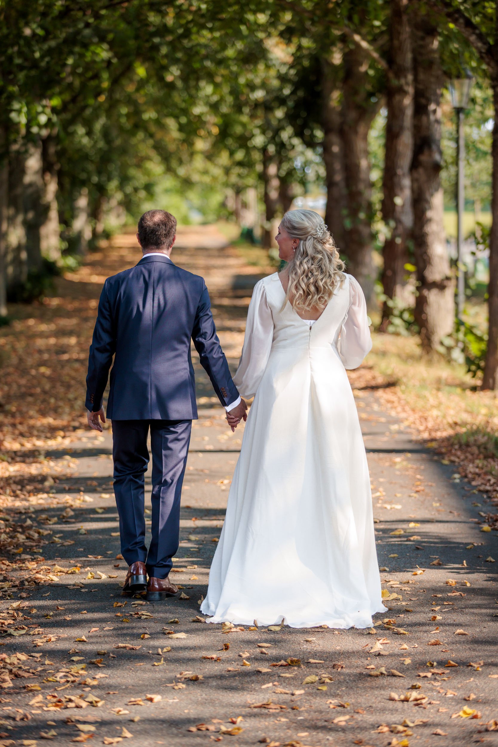 Ett brud- och brudgempaar som gaer hand i hand pa en trädgårdsväg med kastanjelöv. Bruden har en vit brudklanning och berätt hår, medan brudgummen har en marinblå kostym.
