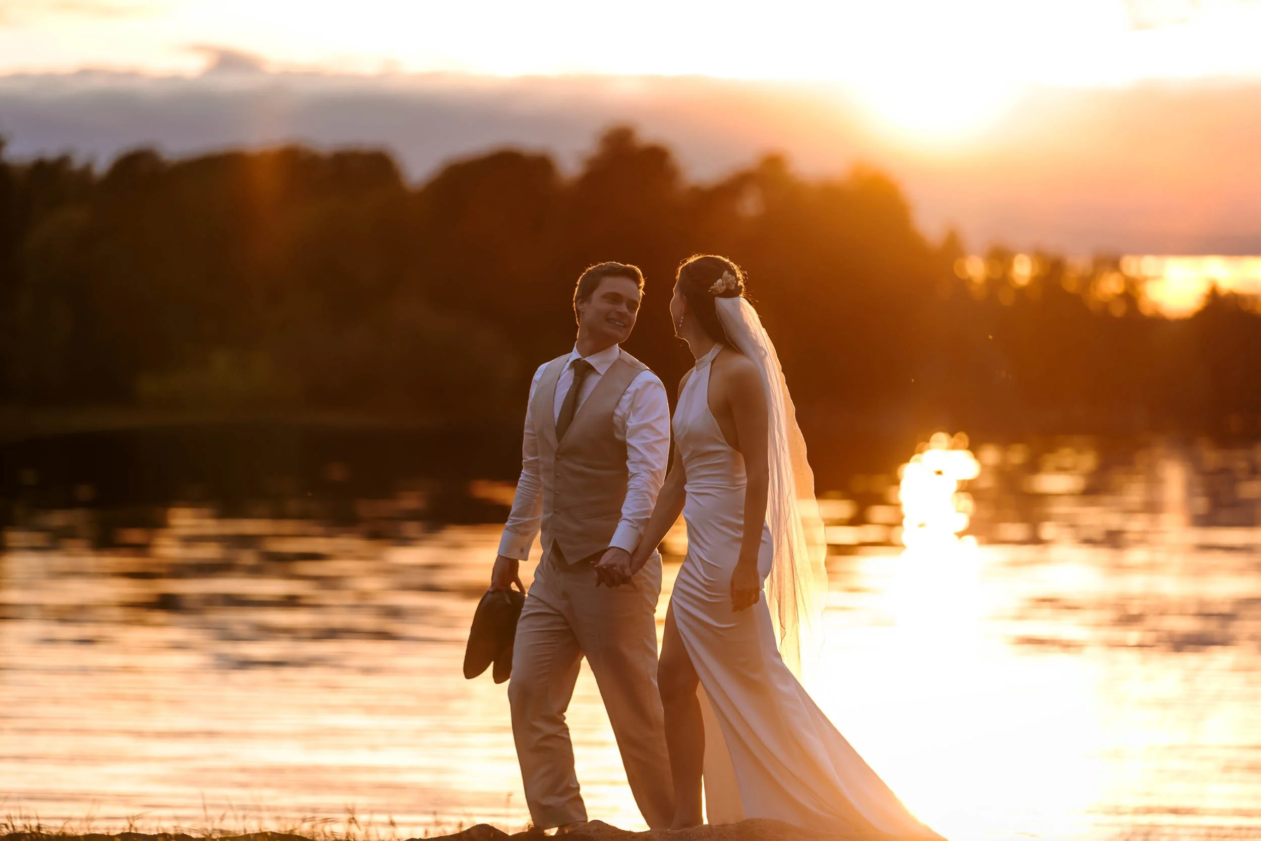 Brudpar håller händer vid sjöstrand i varmt golden hour-ljus med solen speglad i vattnet bakom