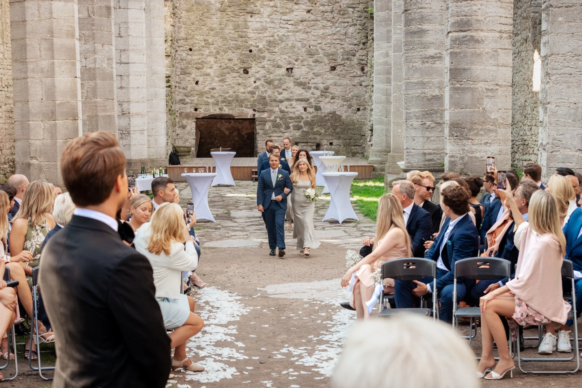 En brud och en brudgum går nerför en kyrkogång inför deras bröllop, omgiven av festgäster som sitter på båda sidor. Ceremonin äger rum i en öppen ruin med höga stenväggar och gräs på marken.